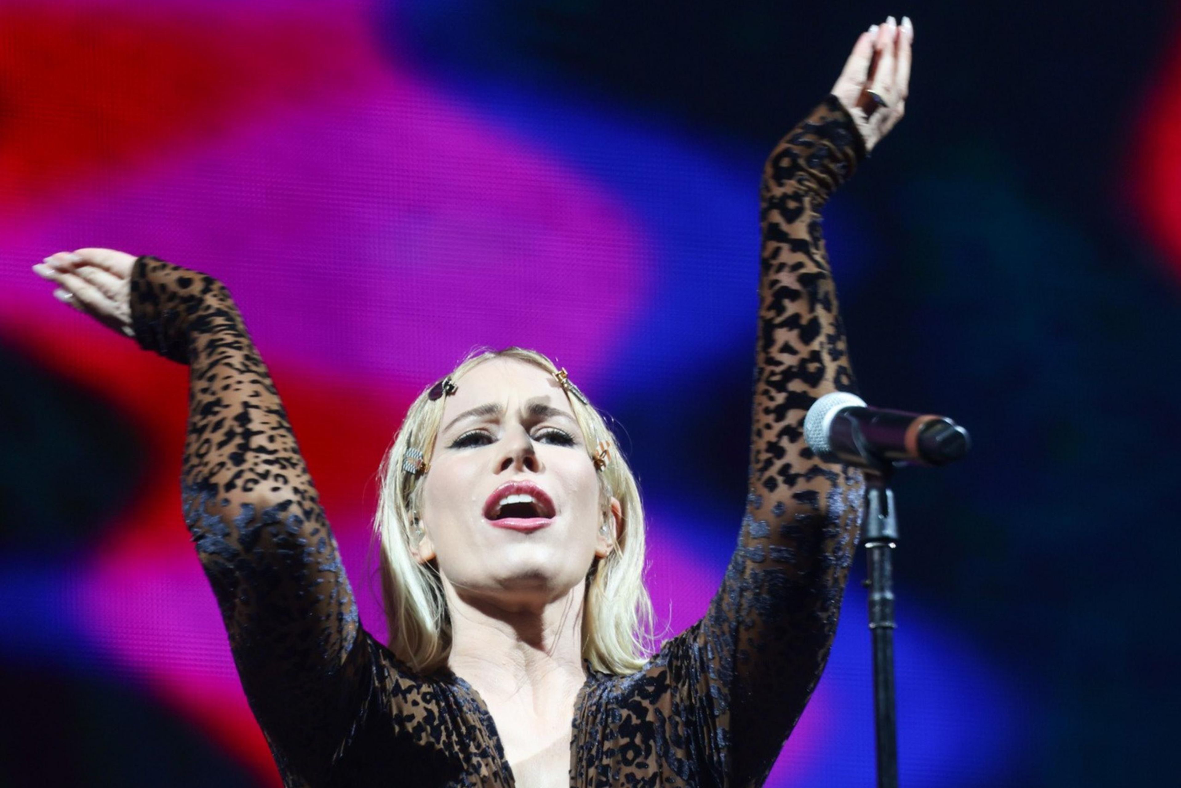 A person wearing a leopard-print jumpsuit performs on stage, with arms raised and a microphone nearby. The background is a colorful, blurred light display.