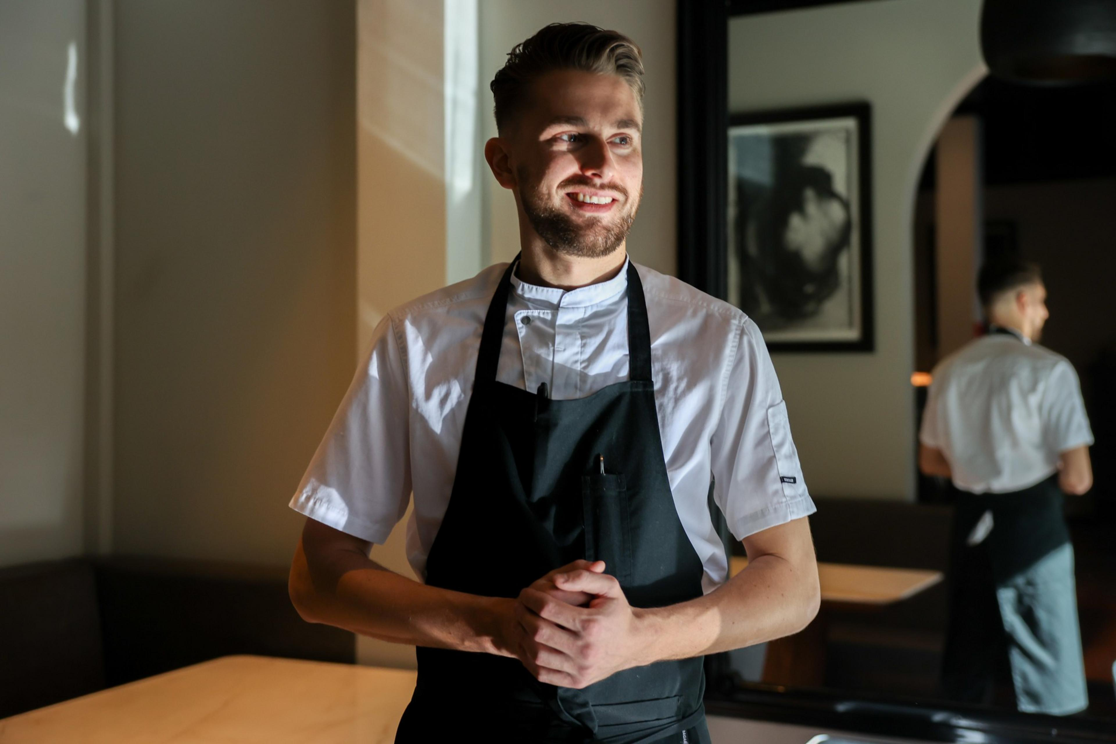 A smiling male chef in a white uniform and black apron stands with clasped hands in a warmly lit restaurant.