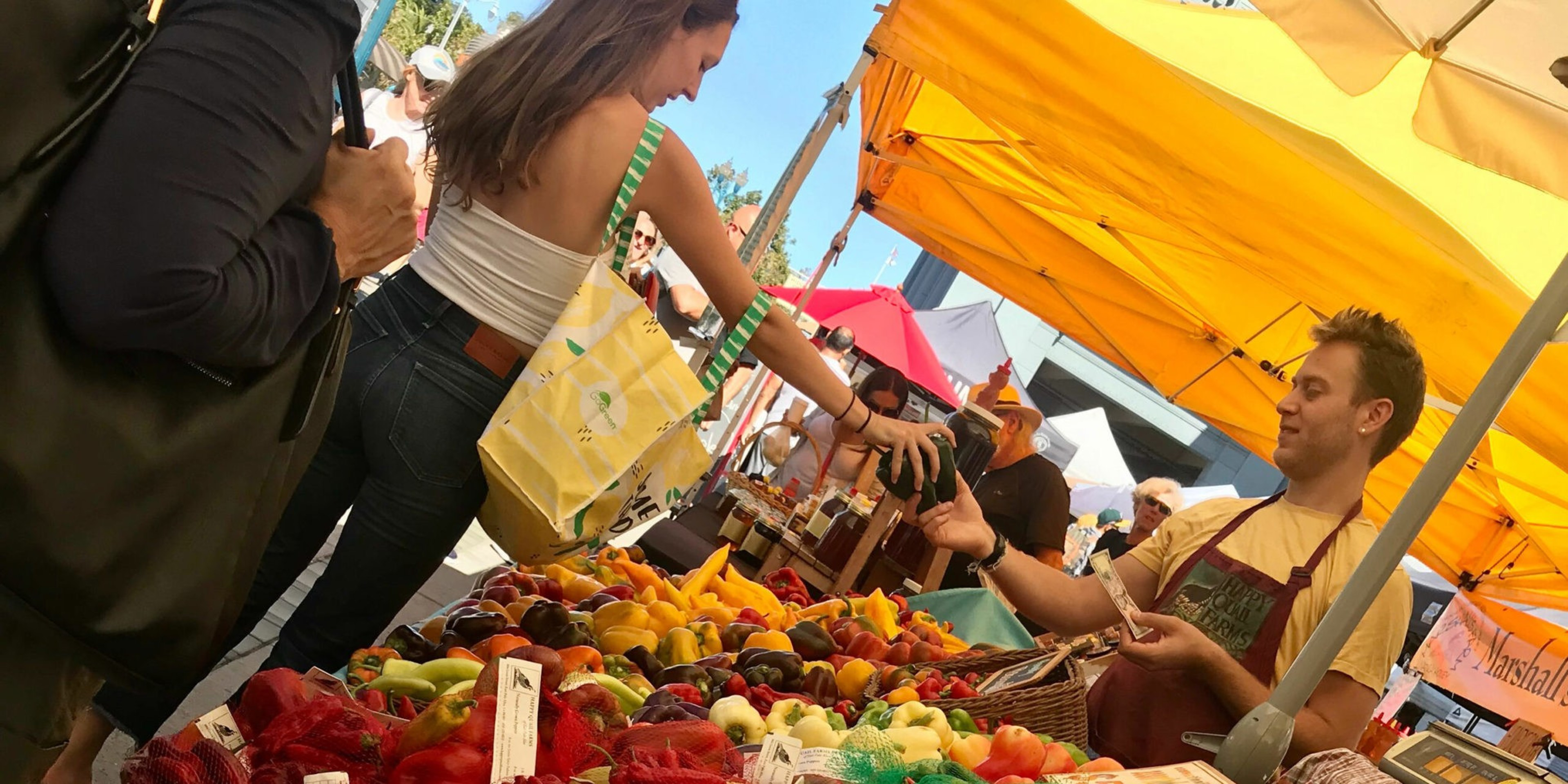 A woman at an outdoor market hands an item to a male vendor under a yellow canopy, with various colorful fruits and vegetables displayed in front of them.