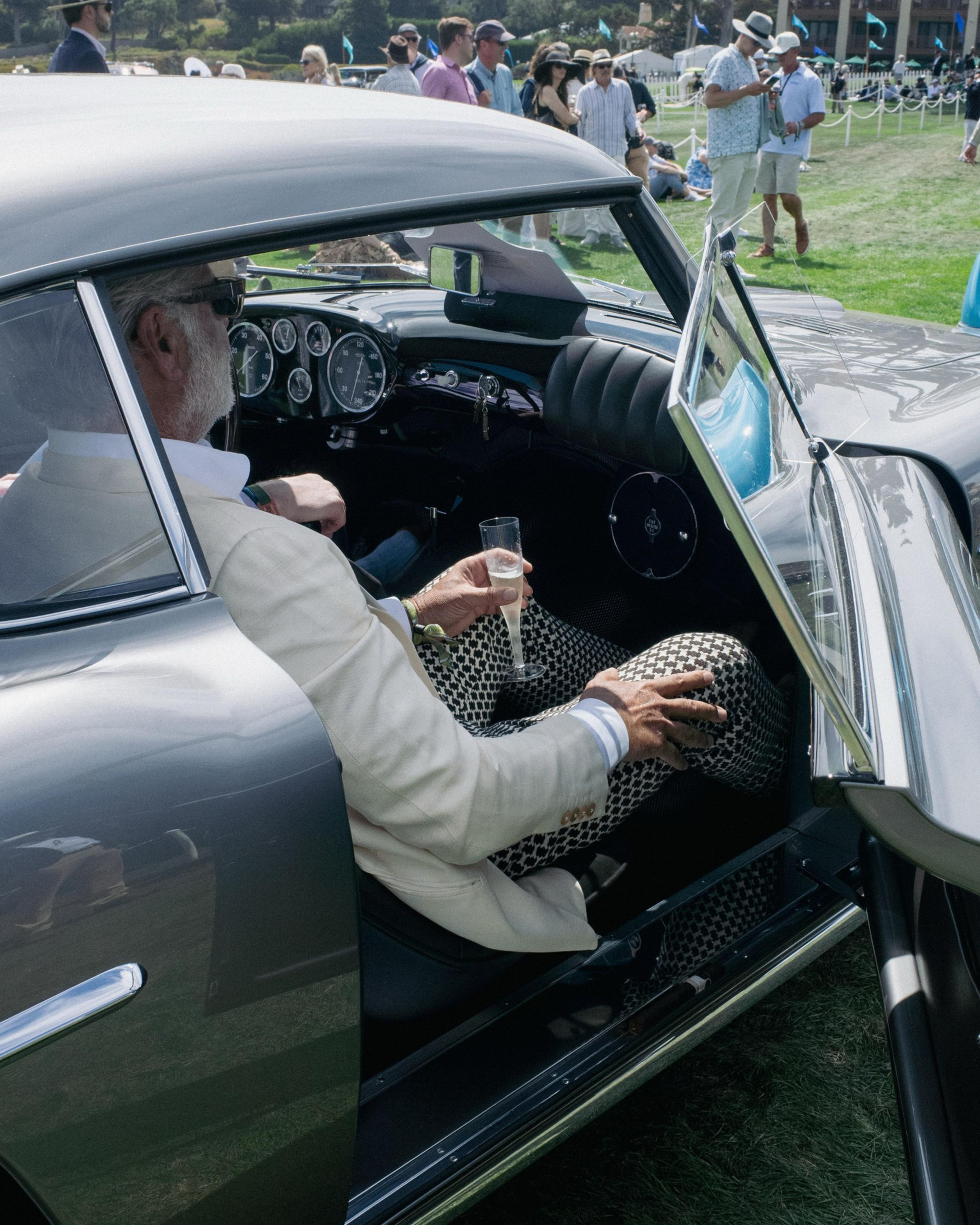An older man in a white blazer and patterned pants sits in a vintage silver car holding a champagne flute, with a crowd visible in the background.