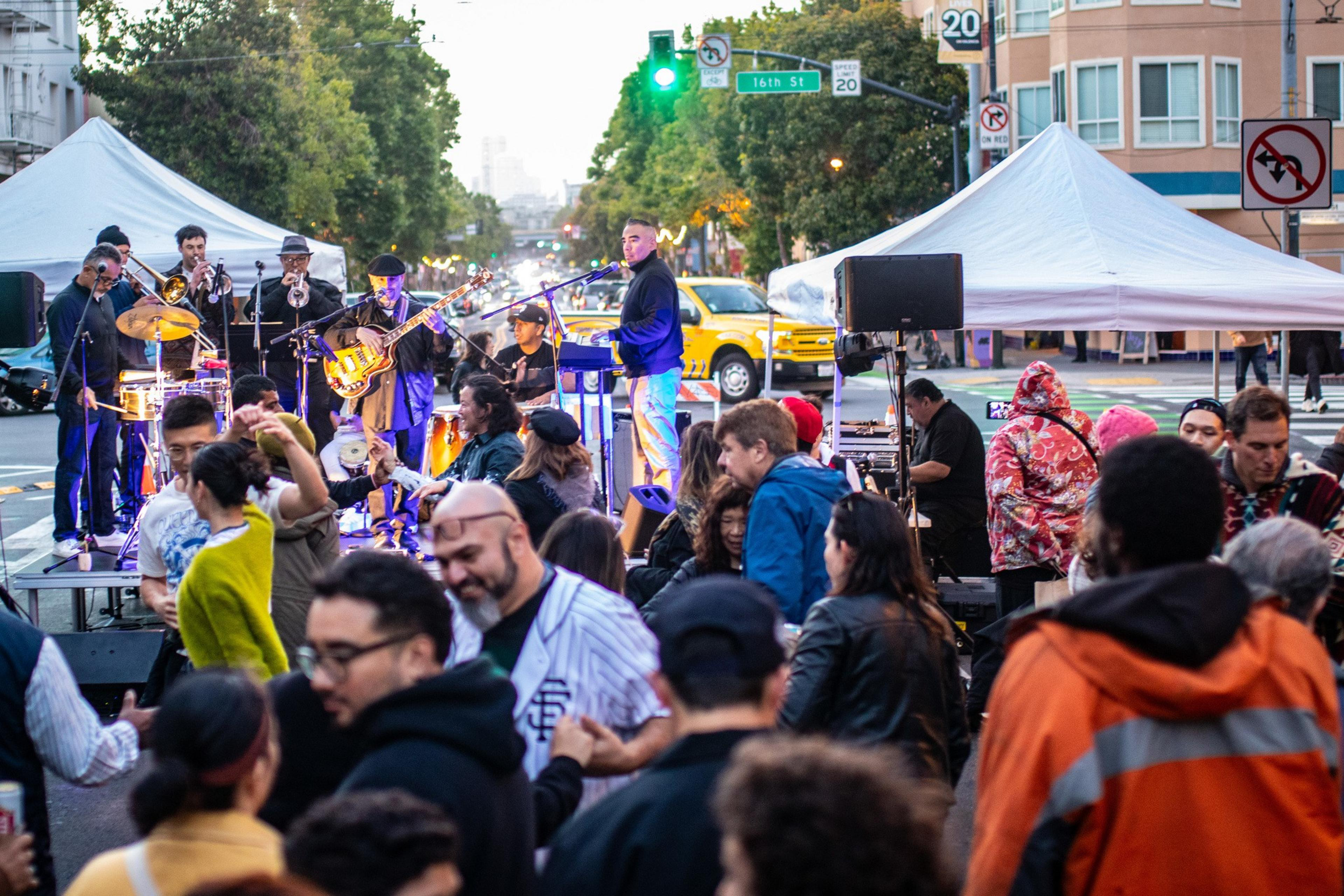 A lively street scene features a band playing under a tent with a crowd dancing and socializing. The background shows a city street with trees and buildings.