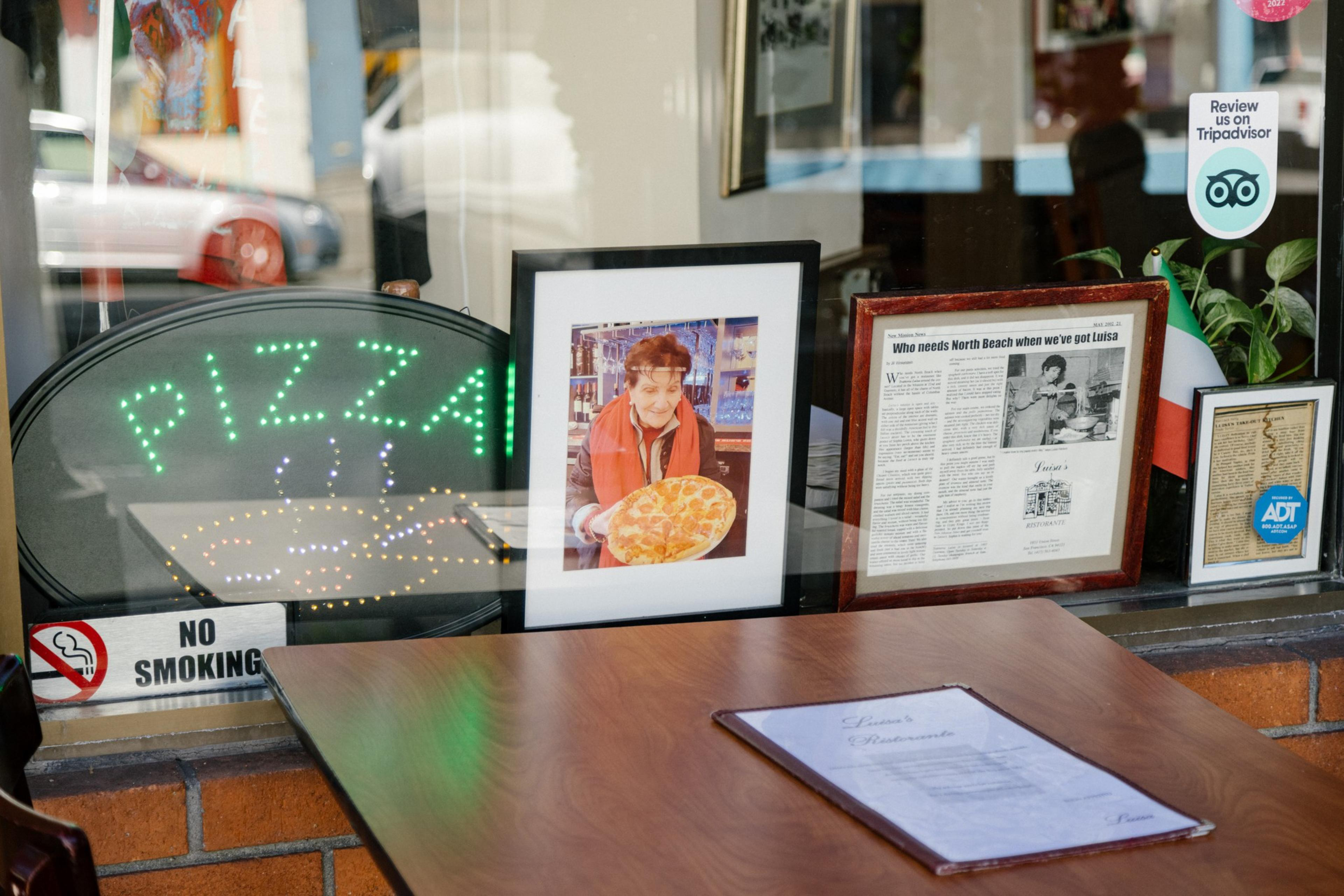 Inside a pizzeria, a neon &quot;PIZZA&quot; sign, articles and a photo of a woman with pizza decorate a window. A menu rests on a table.