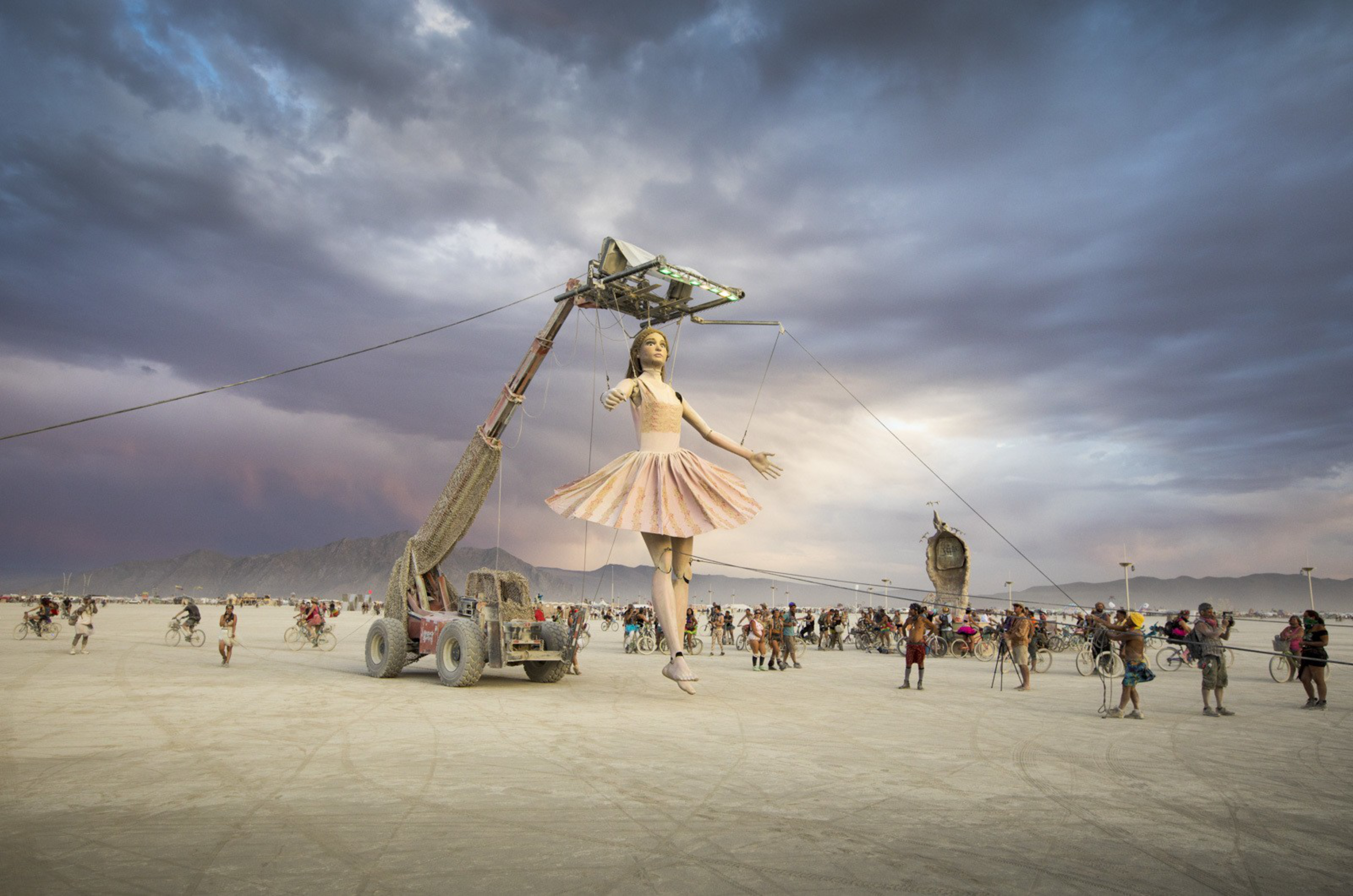 A giant ballerina puppet is suspended by ropes and a mechanical arm in a desert, surrounded by people on bicycles watching it.