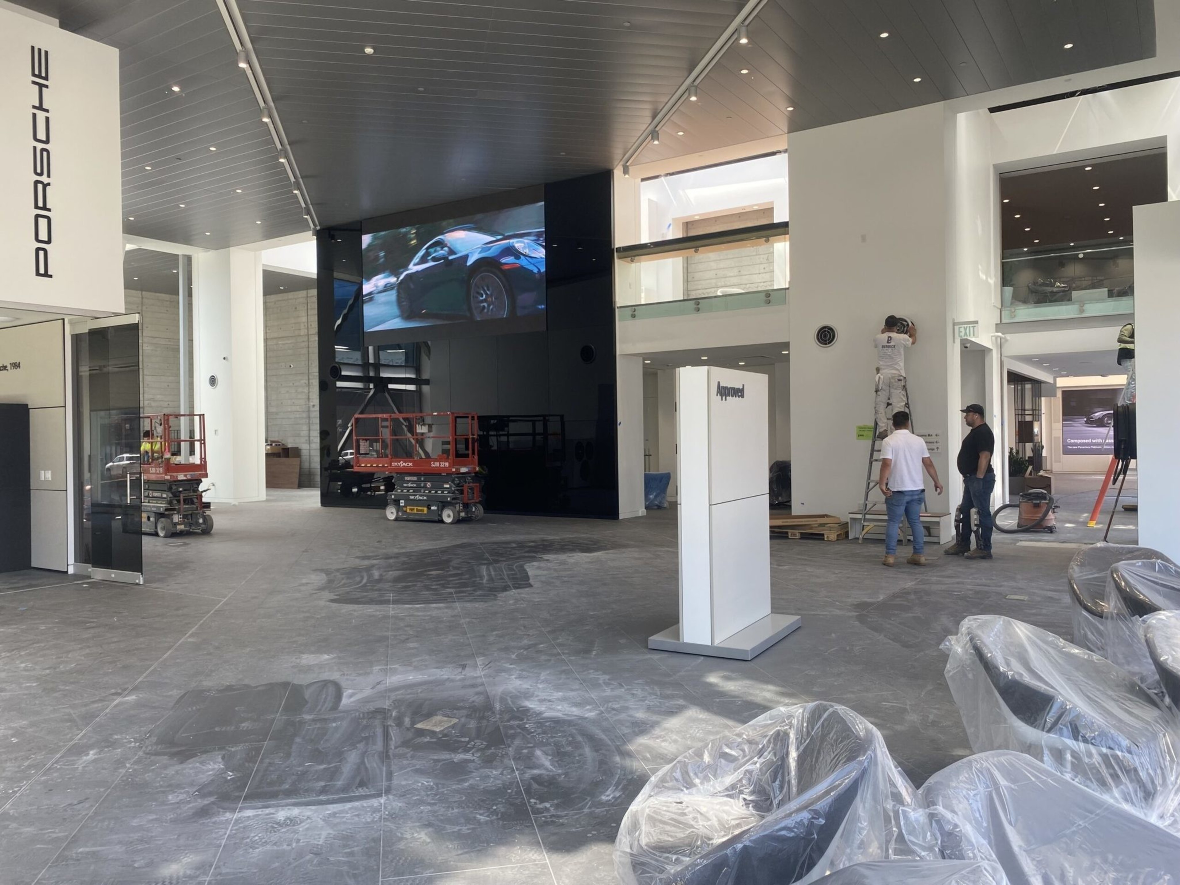 A spacious showroom under construction features a large Porsche sign, workers installing fixtures, covered chairs, and a screen showing a car.