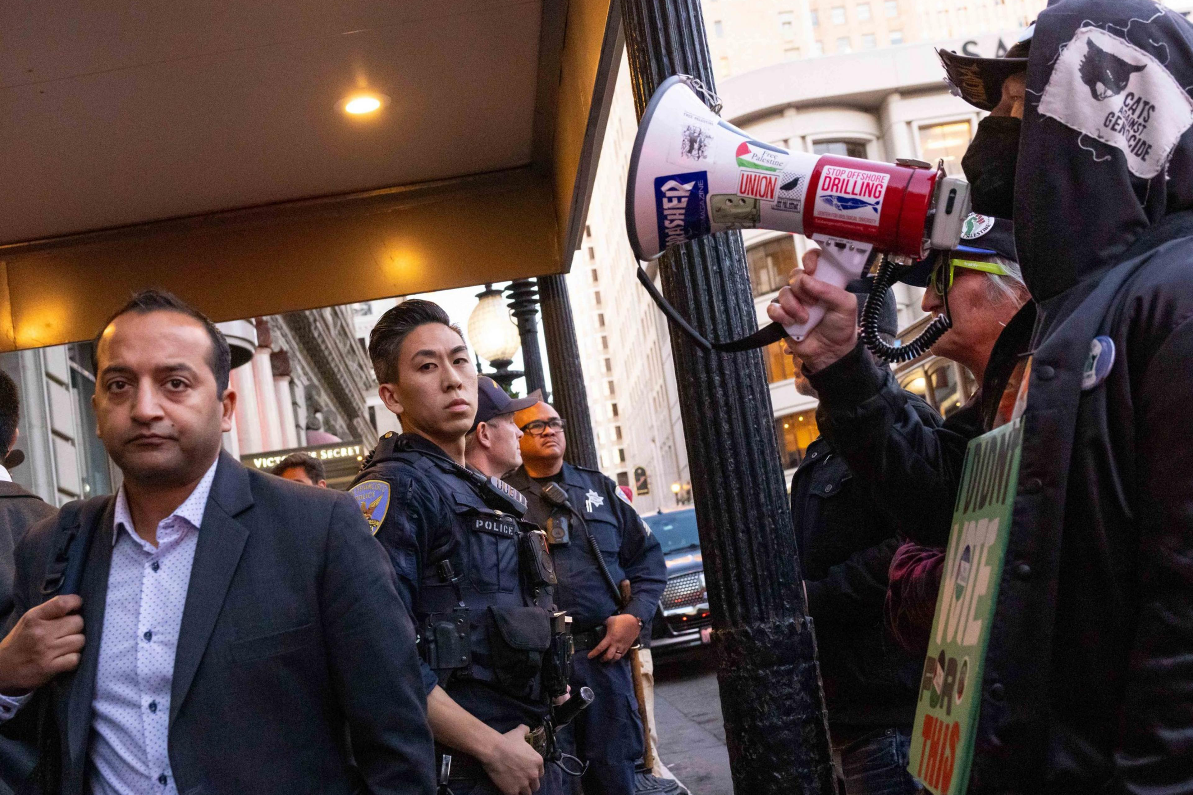 A man walks past police officers on a city street. Nearby, a person holds a megaphone covered in protest stickers, speaking to a small crowd.