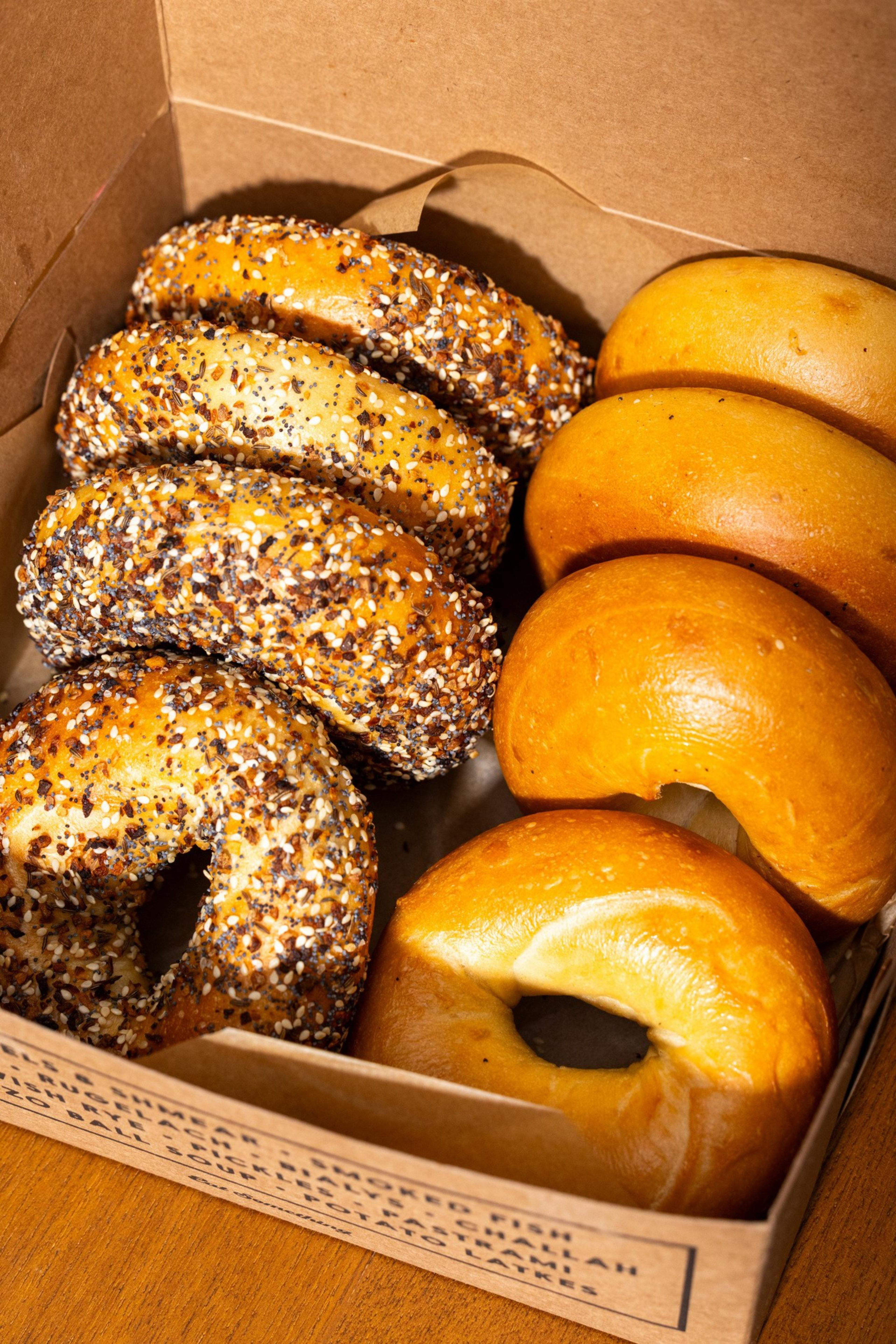 A box contains six bagels; three are covered in seeds and spices, while the other three are plain with a golden-brown crust.