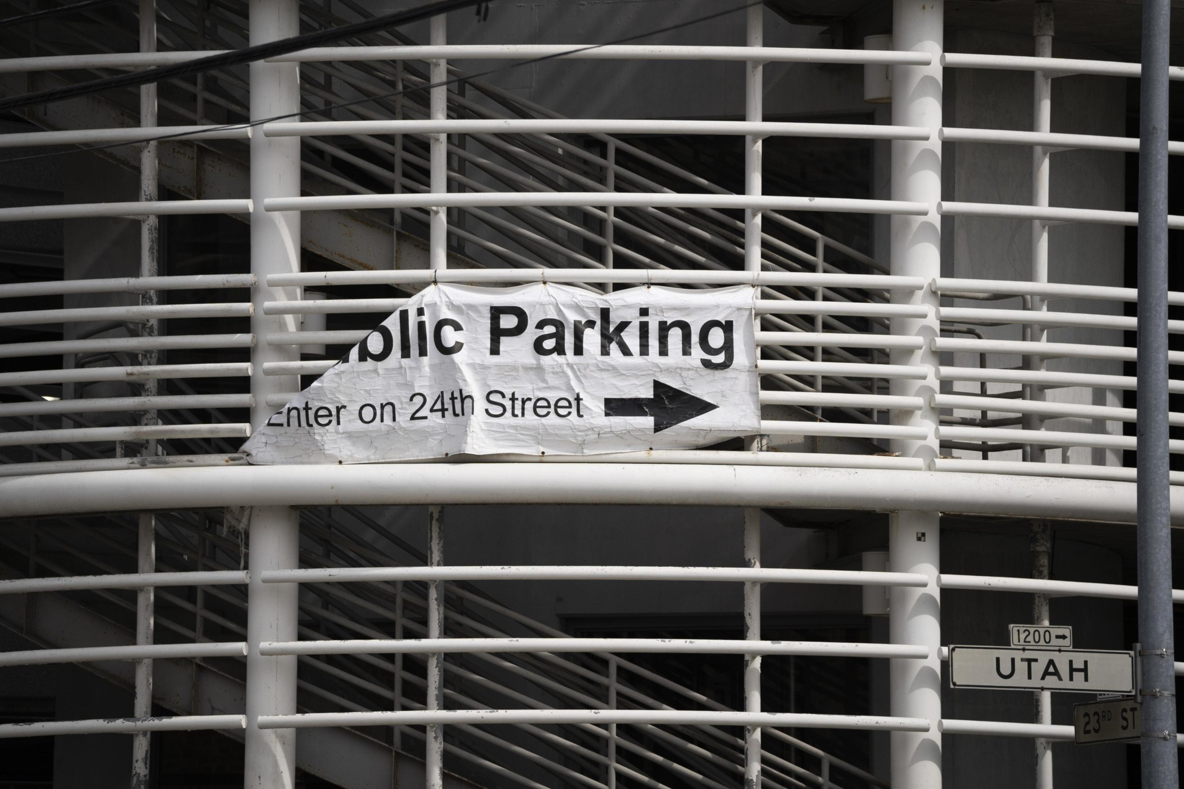 A white sign on a railing reads &quot;Public Parking, Enter on 24th Street&quot; with an arrow. Below are street signs for Utah and 23rd St.