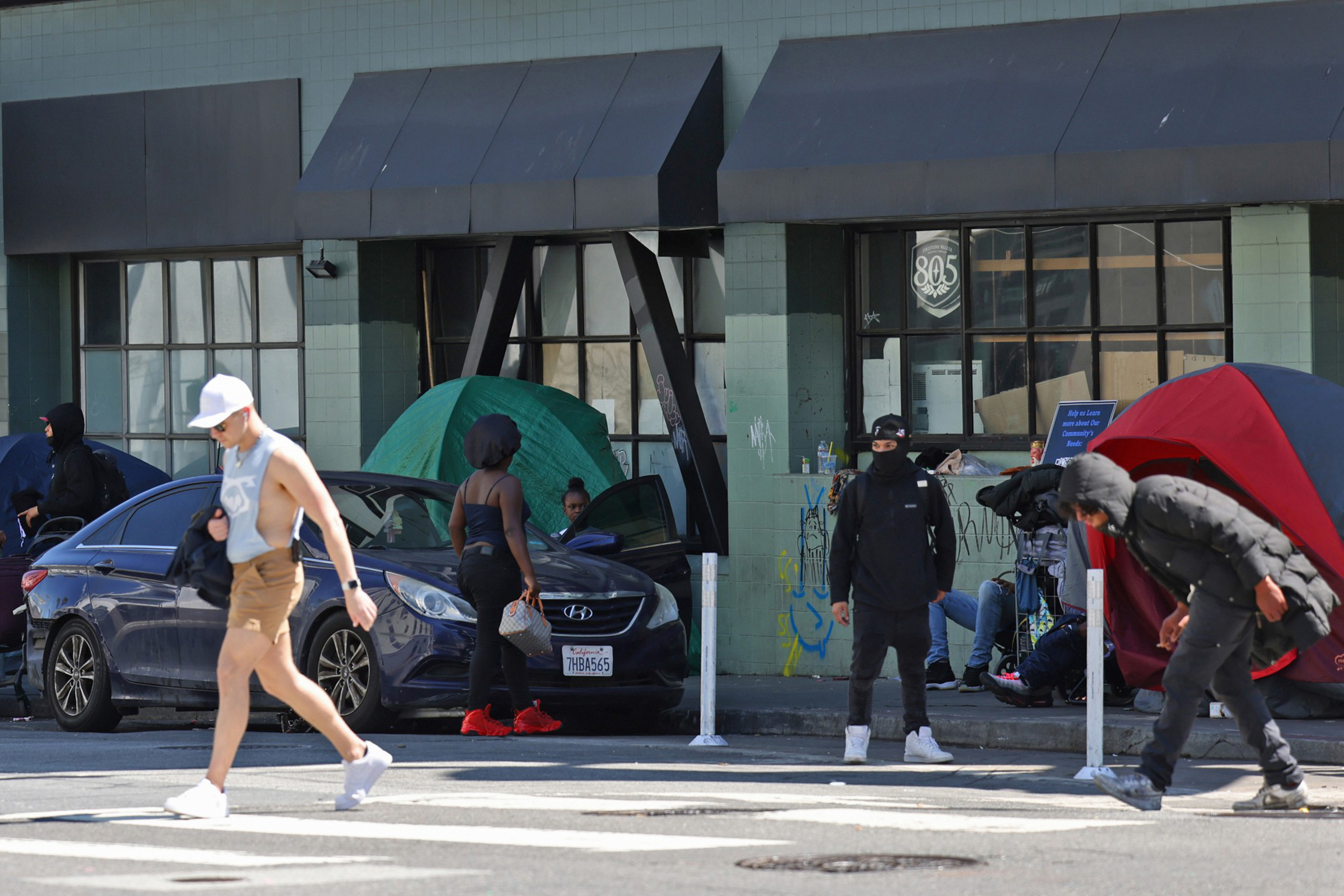 A street view of a homeless encampment along a city street as people walk through the crosswalk on a sunny day.