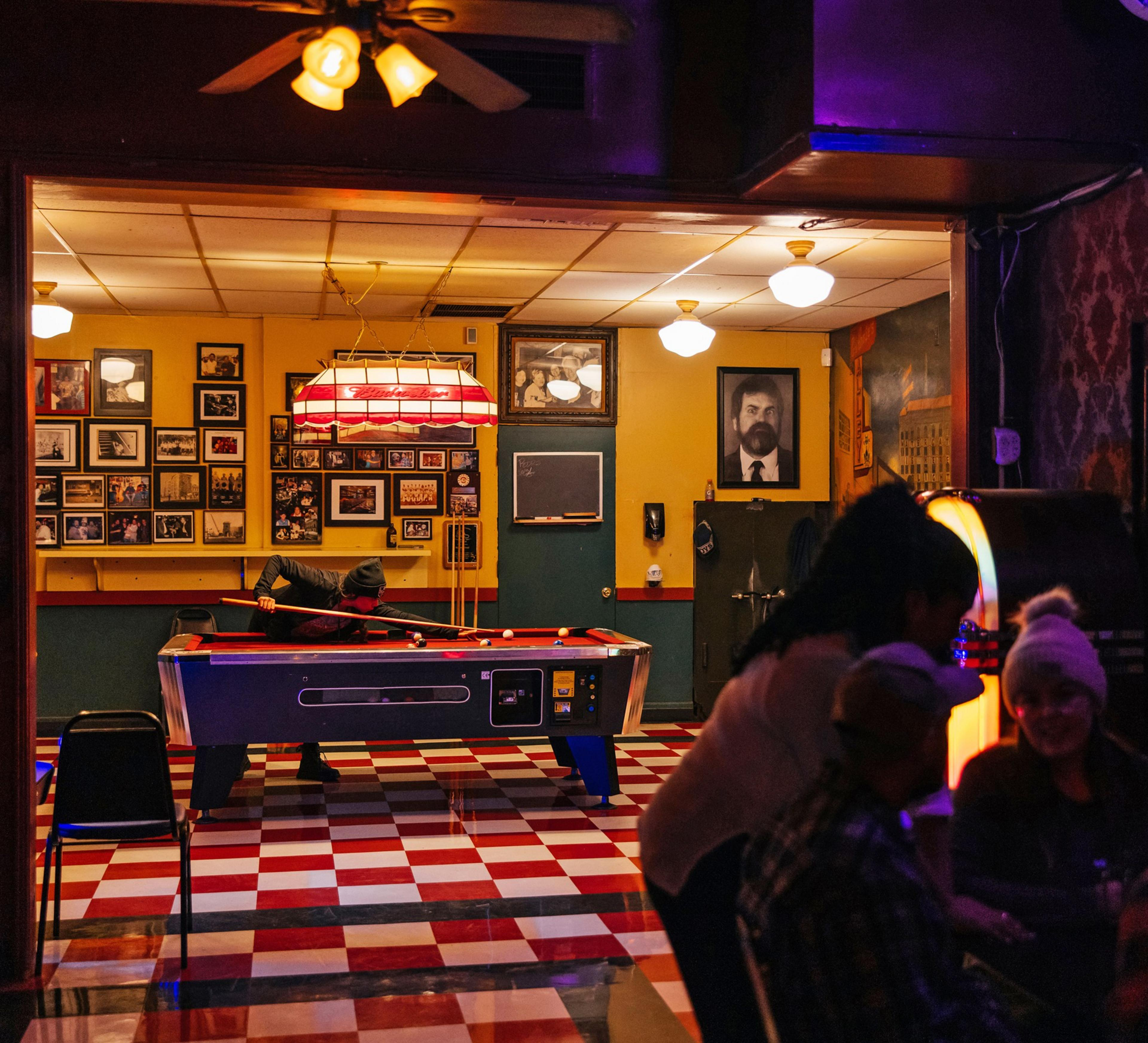 A cozy bar with a red and white checkered floor features a pool table, framed photos on yellow walls, and a jukebox. Patrons are playing pool and socializing.