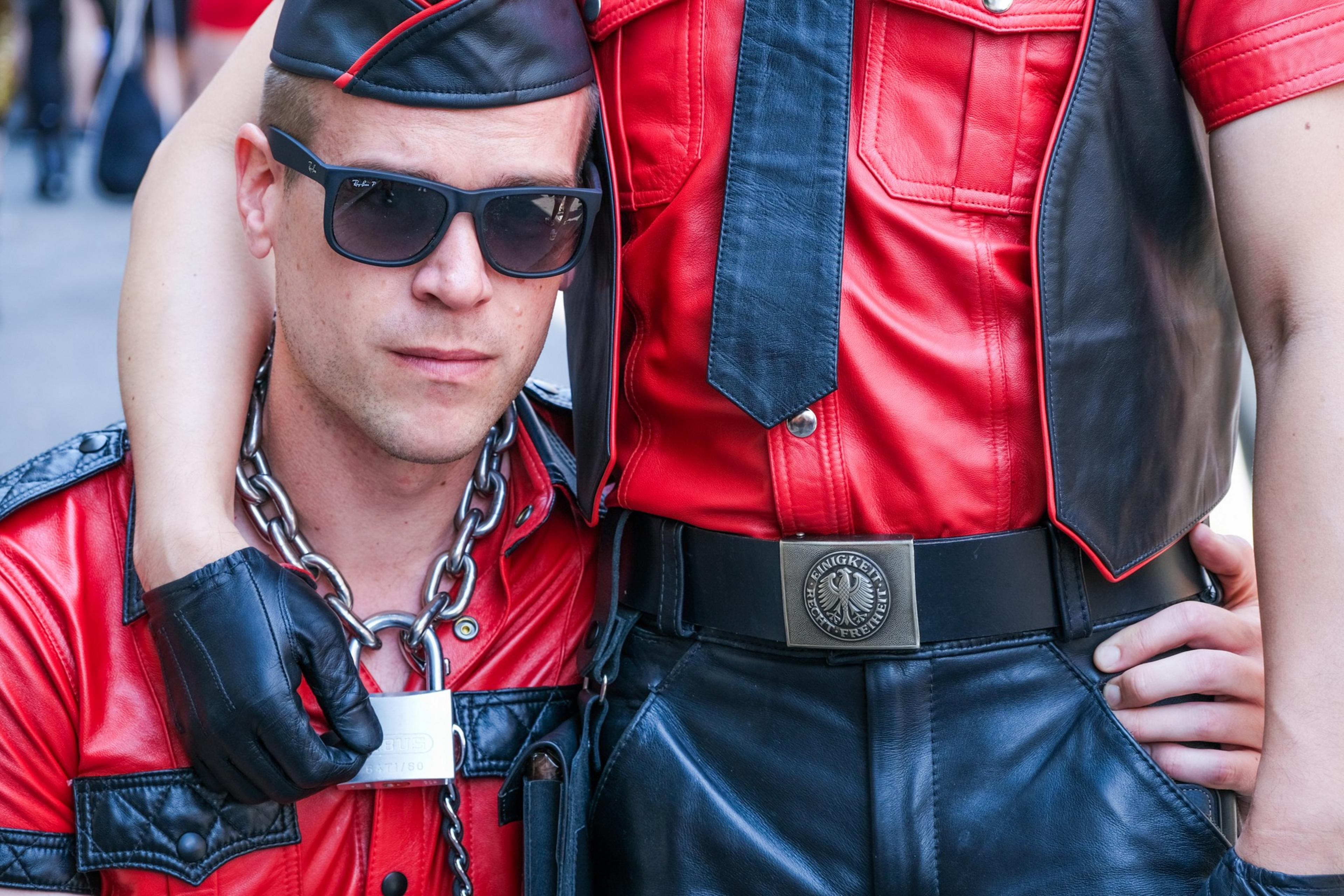 Two people in red and black leather outfits, one wearing sunglasses and a cap, with a chain and lock around the neck, leaning into each other closely.