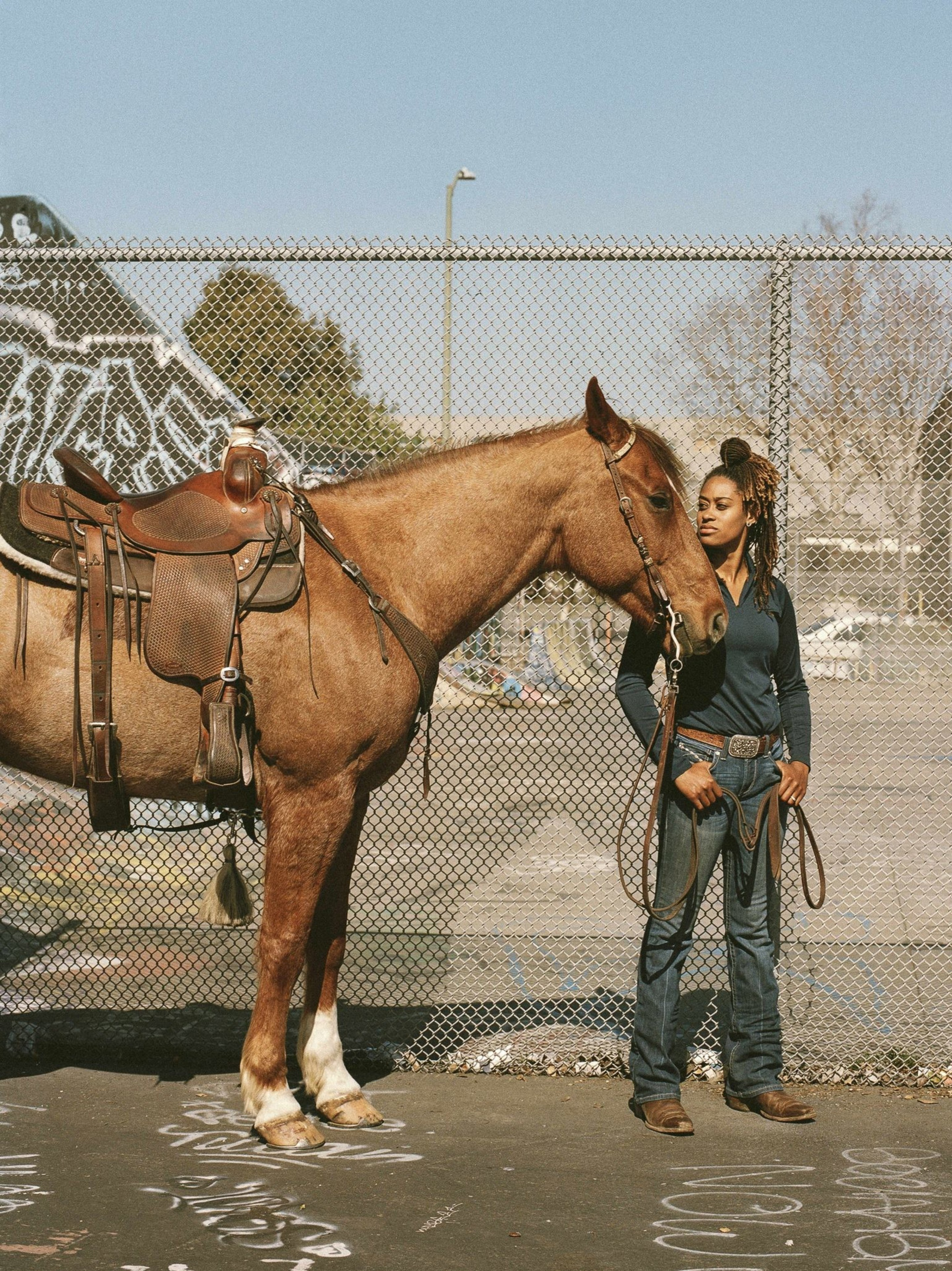 A person stands beside a saddled horse, holding its reins. They are in front of a chain-link fence with some graffiti, under a clear blue sky.