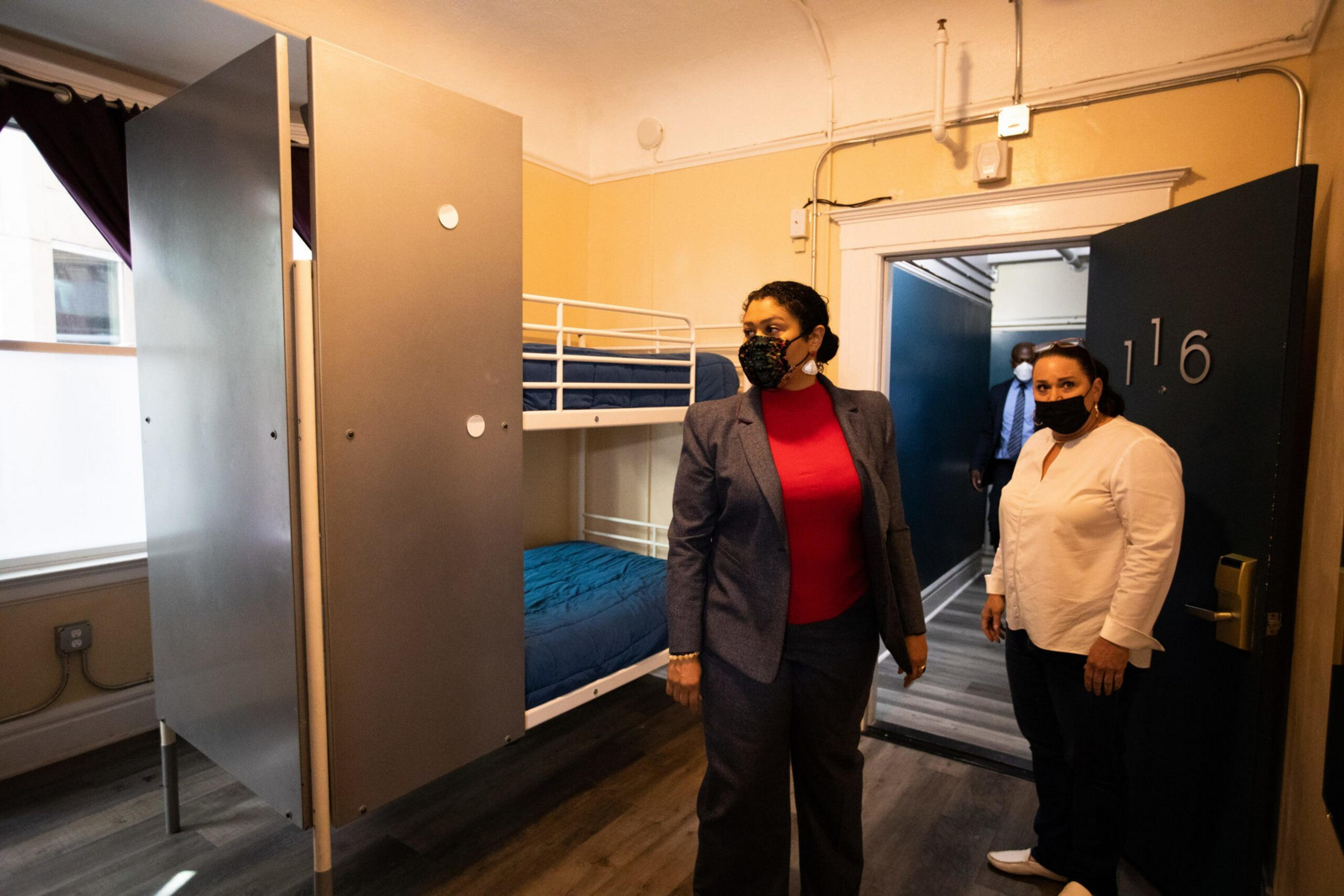 Two women wearing face masks stand inside a small, simple room with a bunk bed and open door numbered 116.