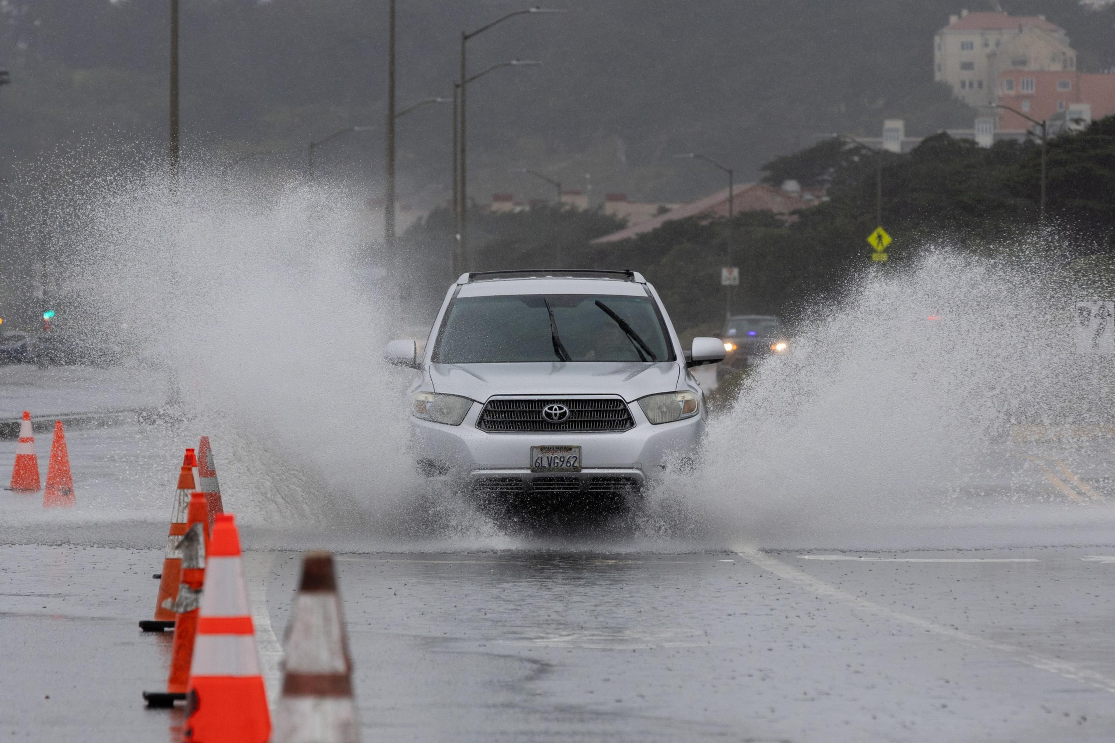 A white SUV splashes through a large puddle on a wet road, with water spraying to the sides.