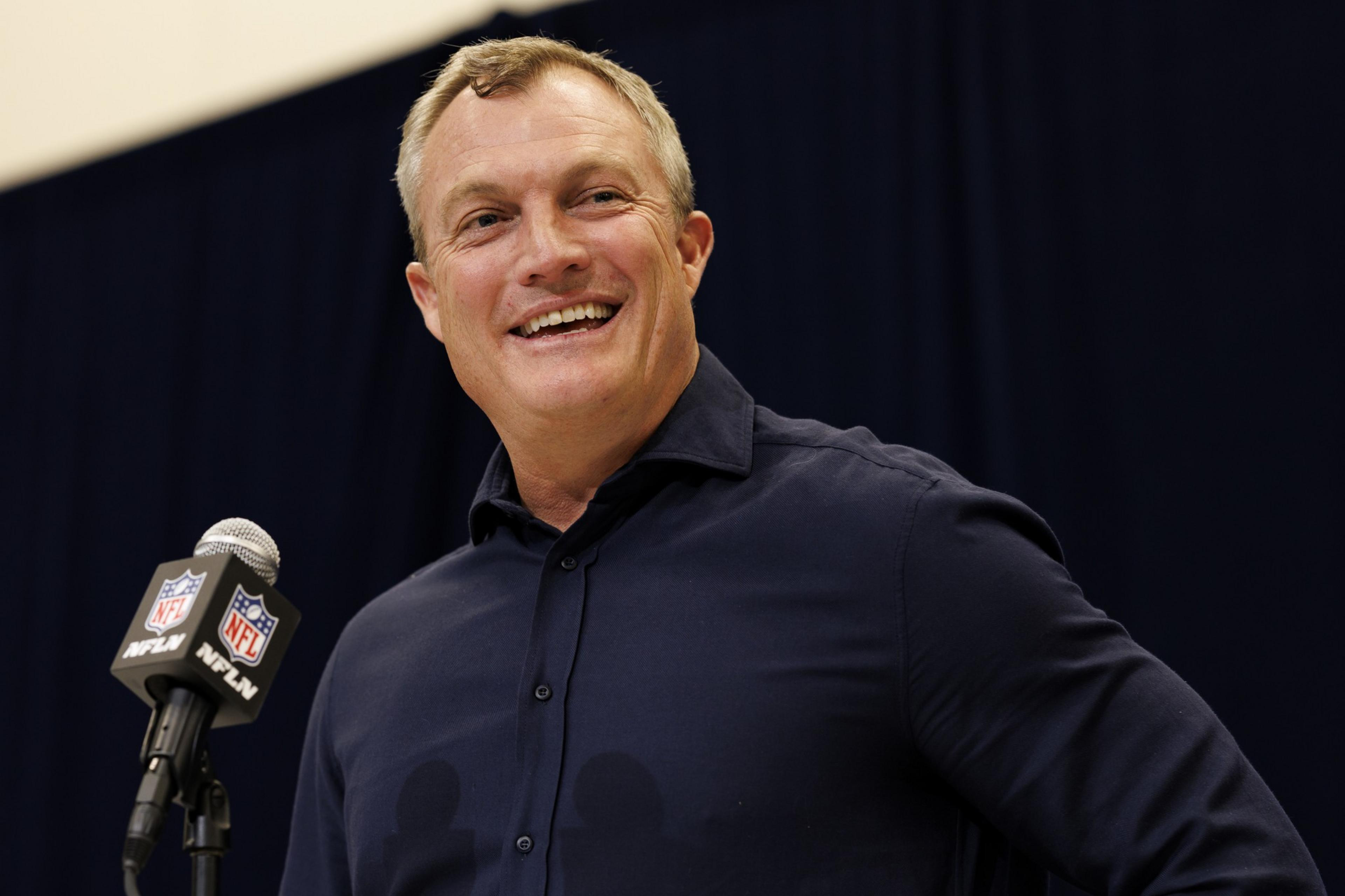 A person in a dark shirt stands smiling beside a microphone with the NFL logo. The background is dark.