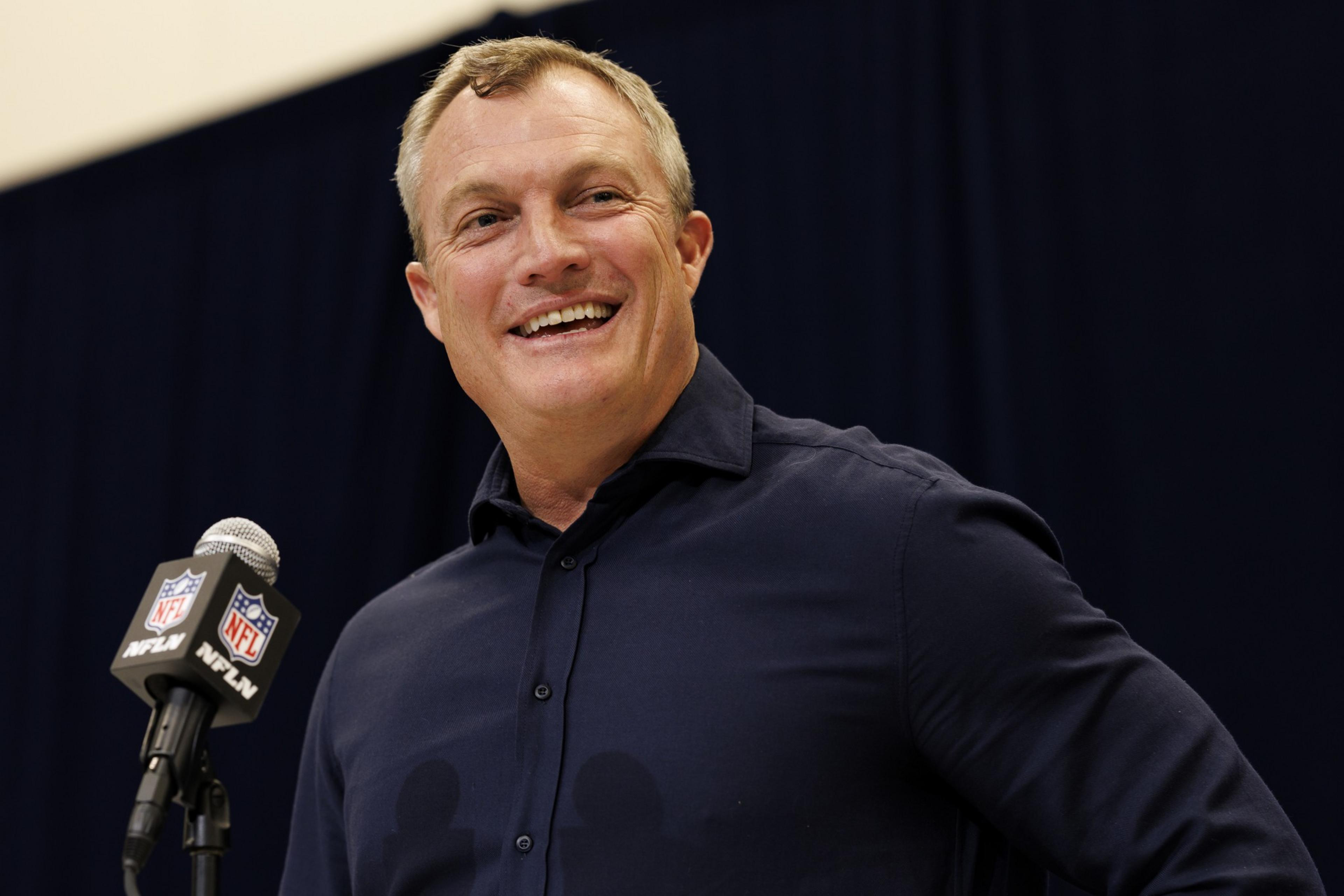 A person in a dark shirt stands smiling beside a microphone with the NFL logo. The background is dark.