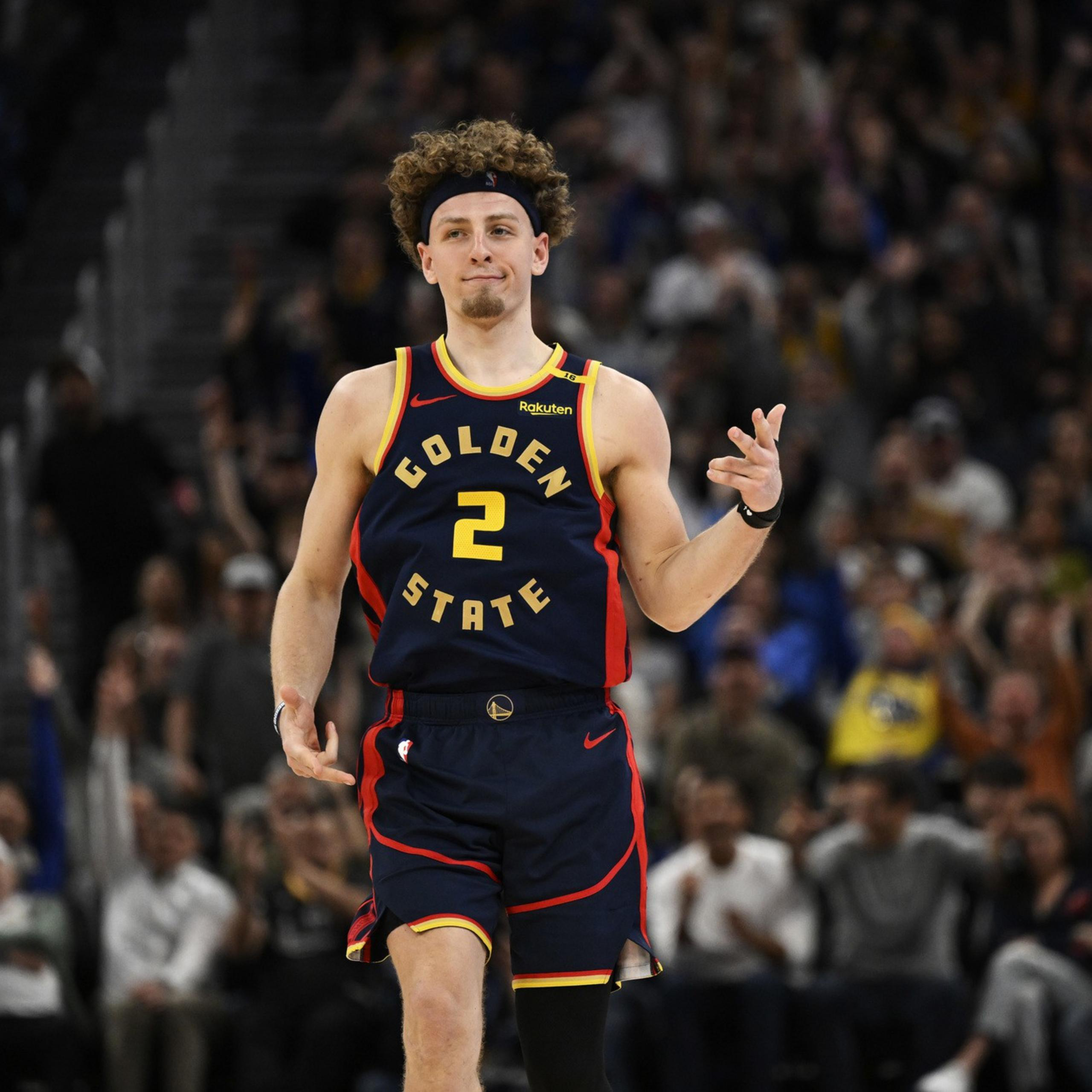 A basketball player wearing a Golden State jersey with the number 2 is making a hand gesture, possibly celebrating. A crowd in the background looks cheering.