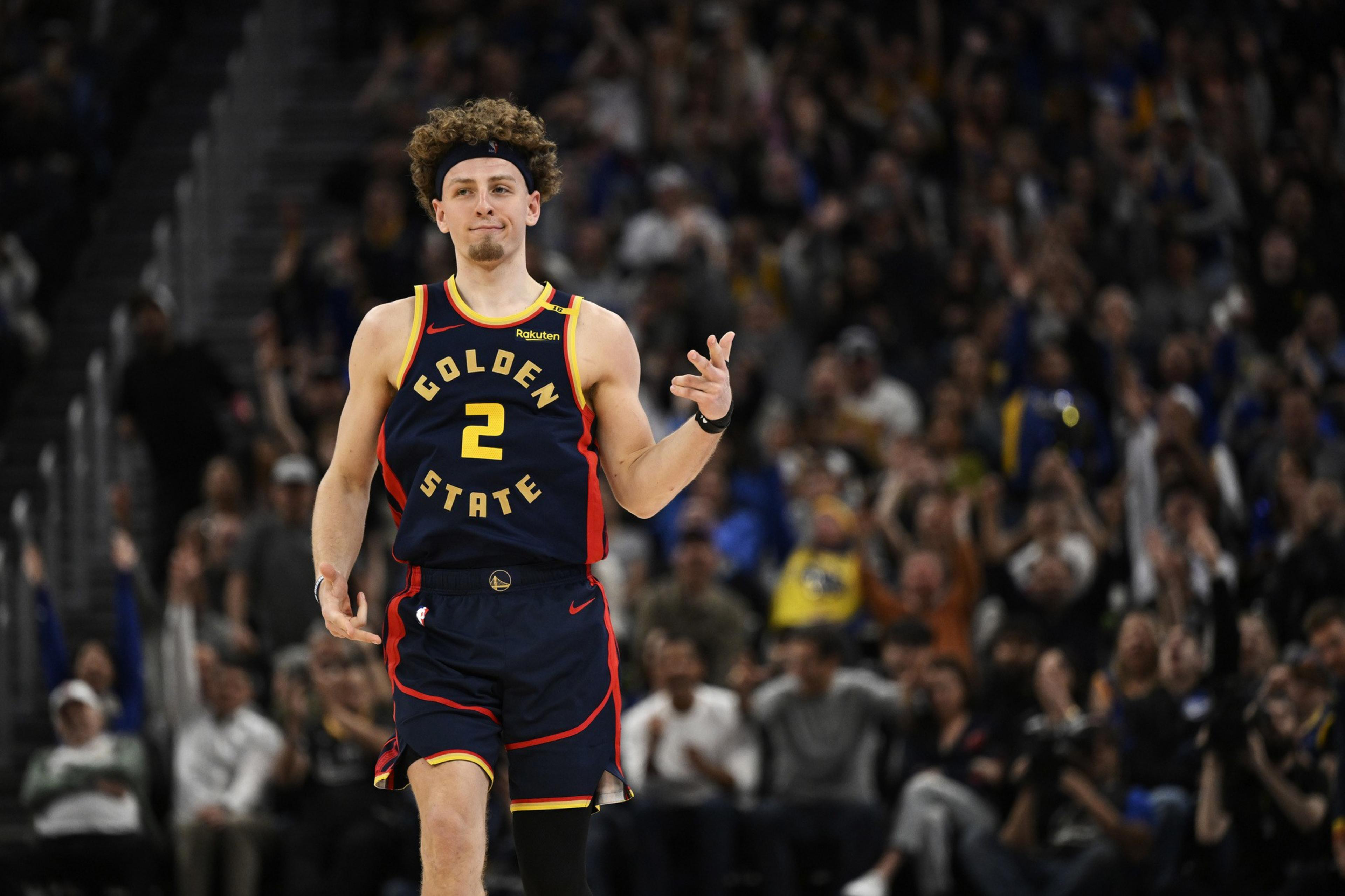 A basketball player wearing a Golden State jersey with the number 2 is making a hand gesture, possibly celebrating. A crowd in the background looks cheering.