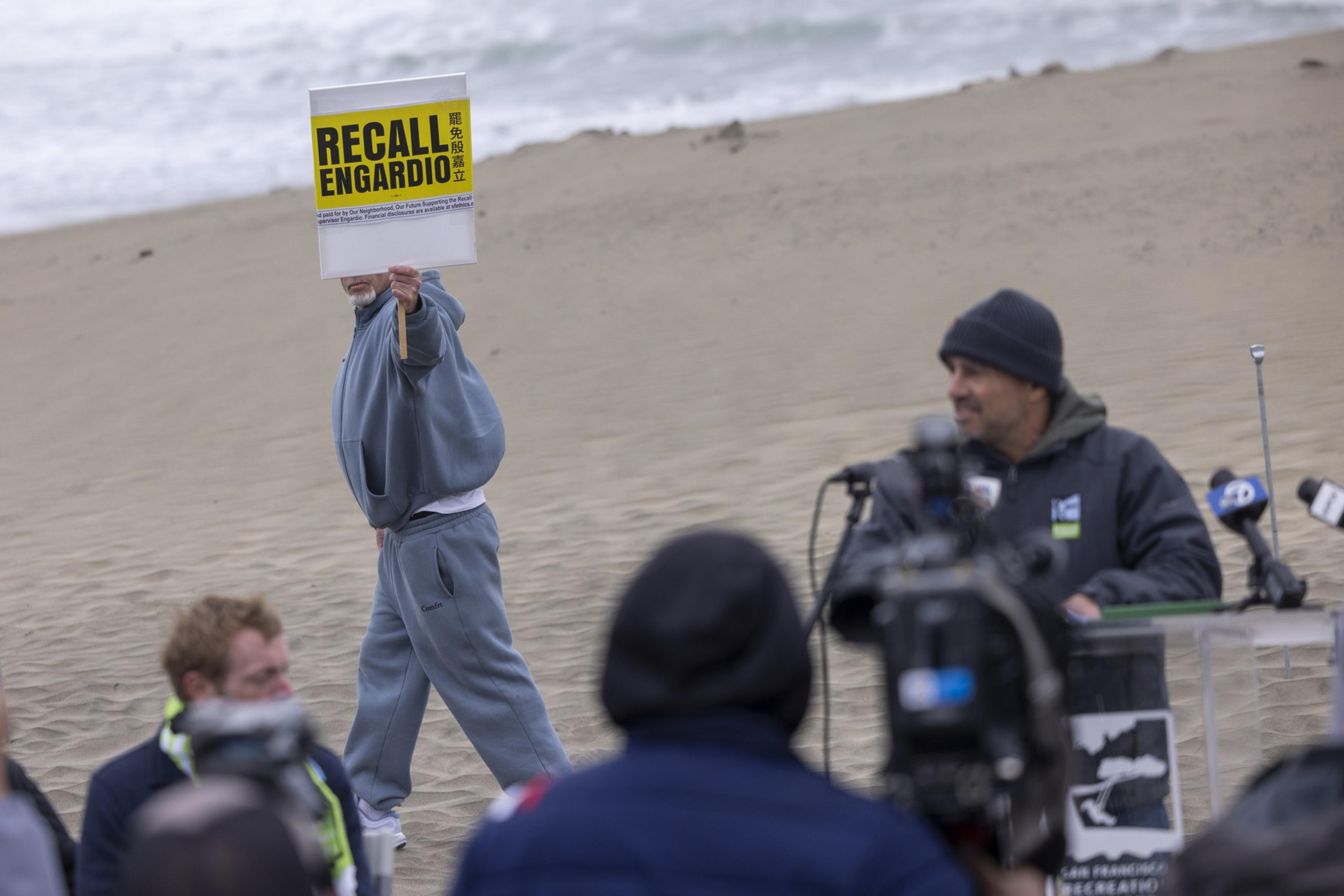 A person in blue holds a "RECALL ENGARDIO" sign on a beach, while several people and cameras focus on a speaker at a podium nearby.