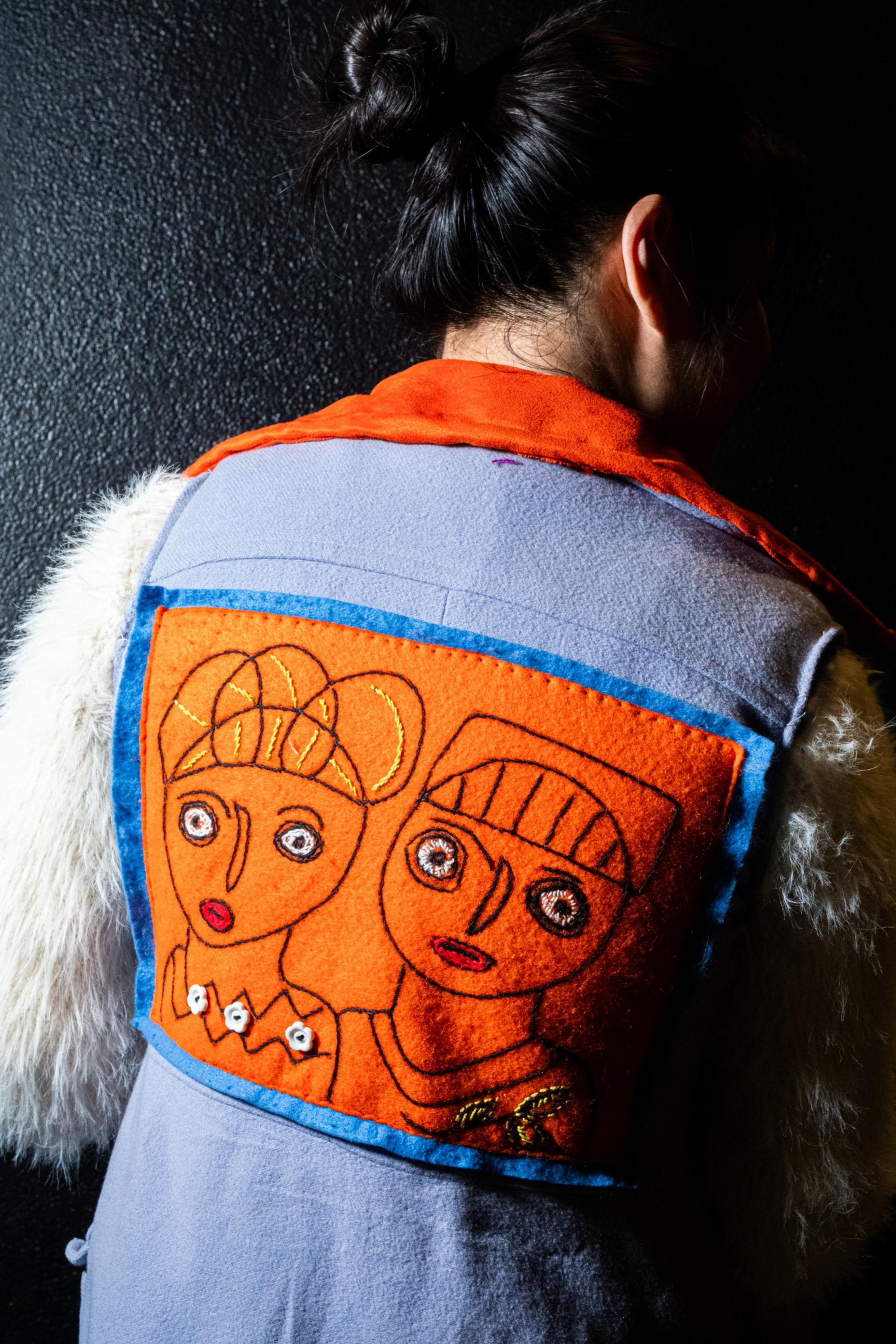 A person with a dark hair bun wears a colorful jacket featuring a stitched patch of abstract human figures with expressive faces on the back.