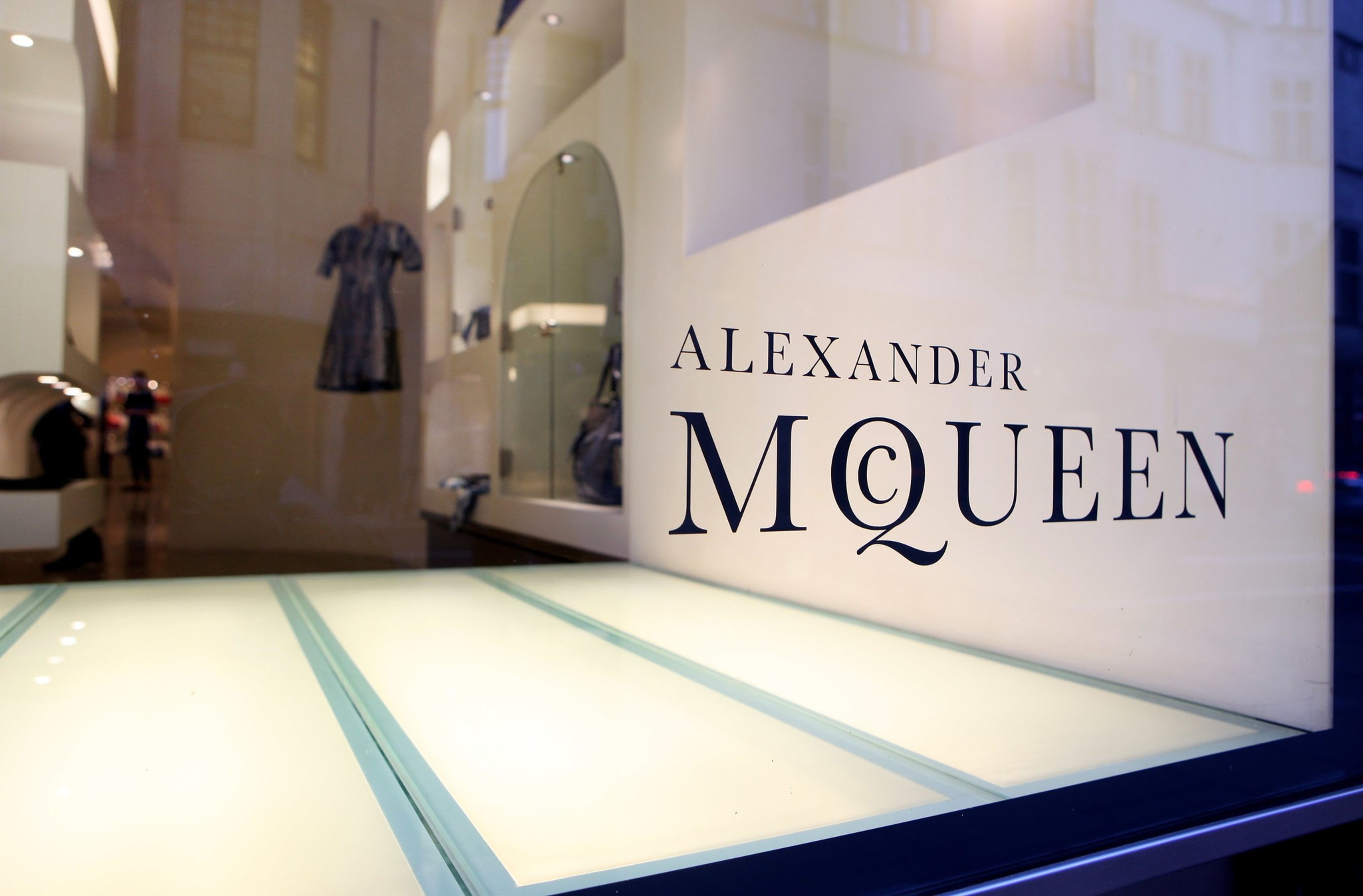 A store window displays the text &quot;ALEXANDER McQUEEN&quot; in bold letters. Inside, a single dress is visible, hanging centrally in a brightly lit space.