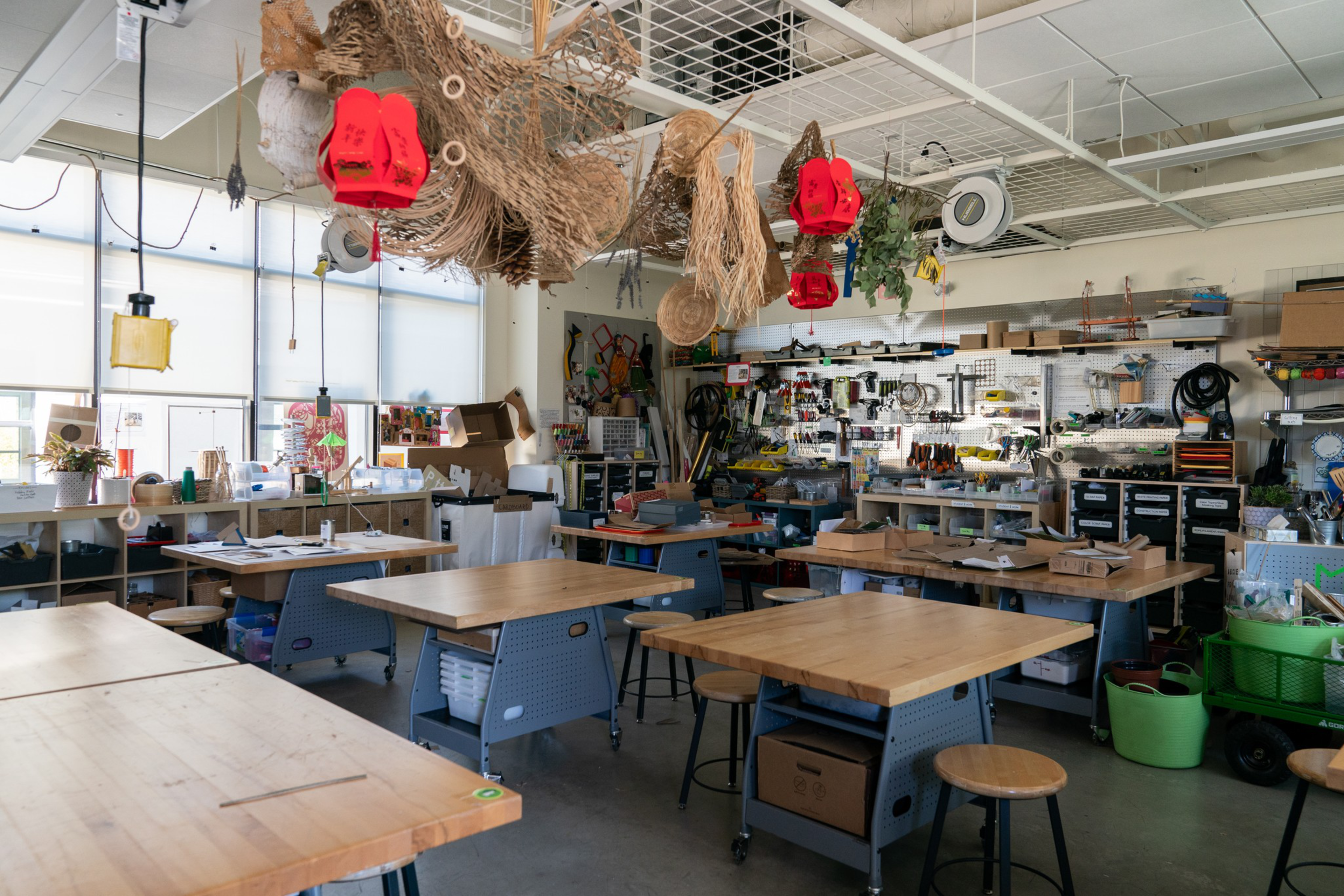 The image depicts a bright, organized workshop with wooden tables, stools, hanging crafts, tools on pegboards, and various storage bins and shelves lining the walls.