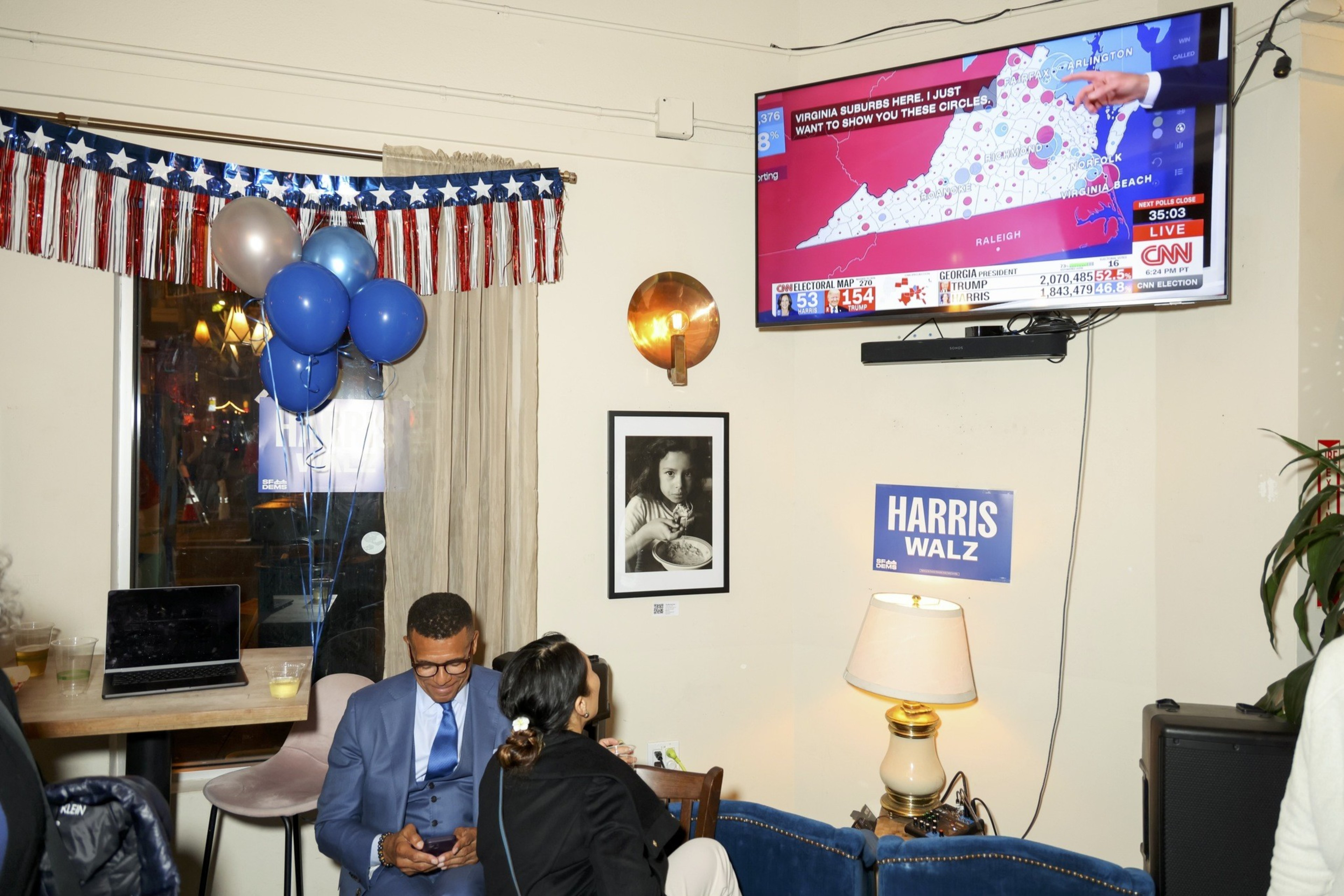 The image depicts an election watch party with people seated, balloons, a CNN map on TV, and campaign materials including a &quot;HARRIS WALZ&quot; sign.