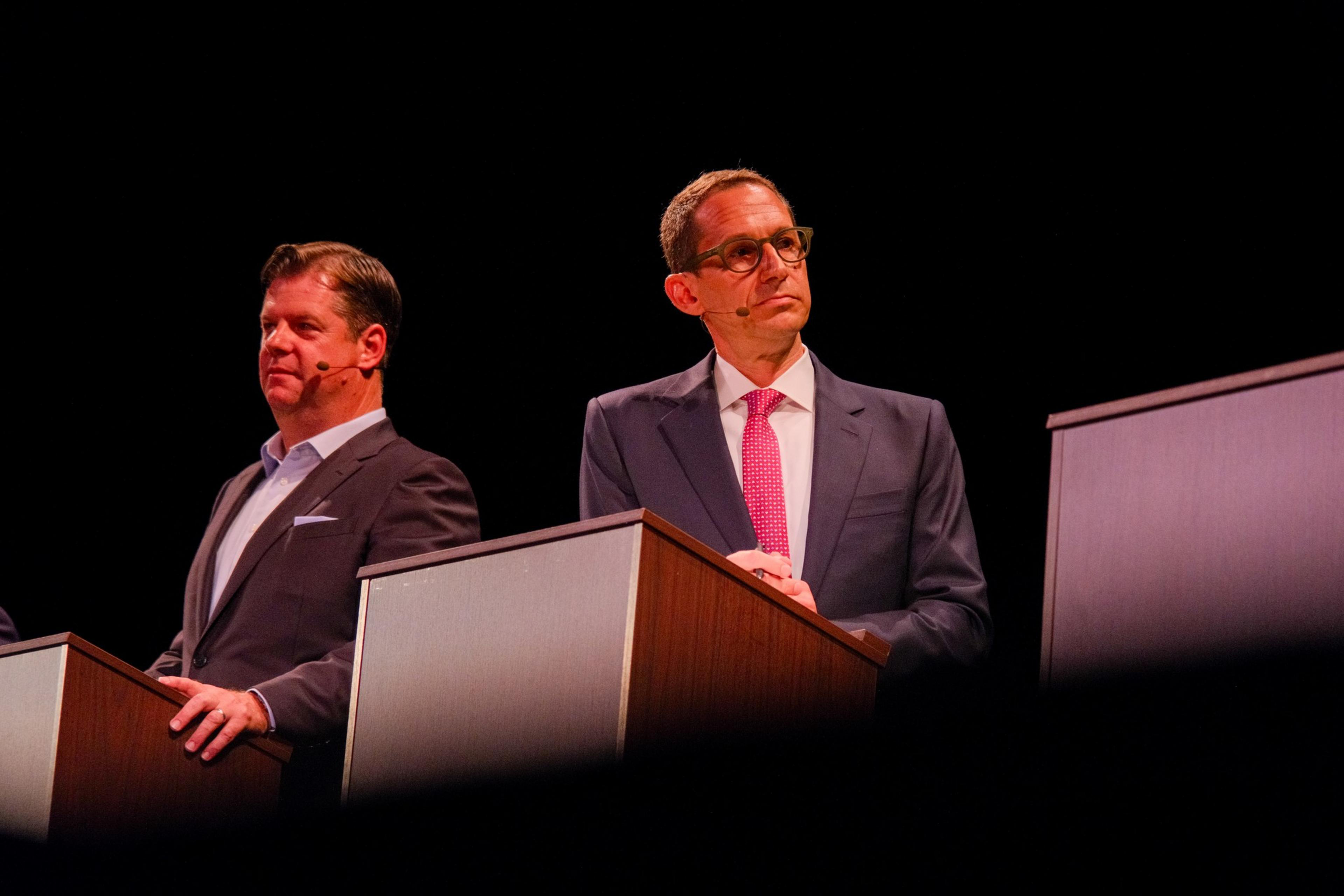 Two men in suits stand behind podiums on a dark stage, wearing microphones on their faces. One man wears glasses and a pink tie; the other man wears a light blue shirt.