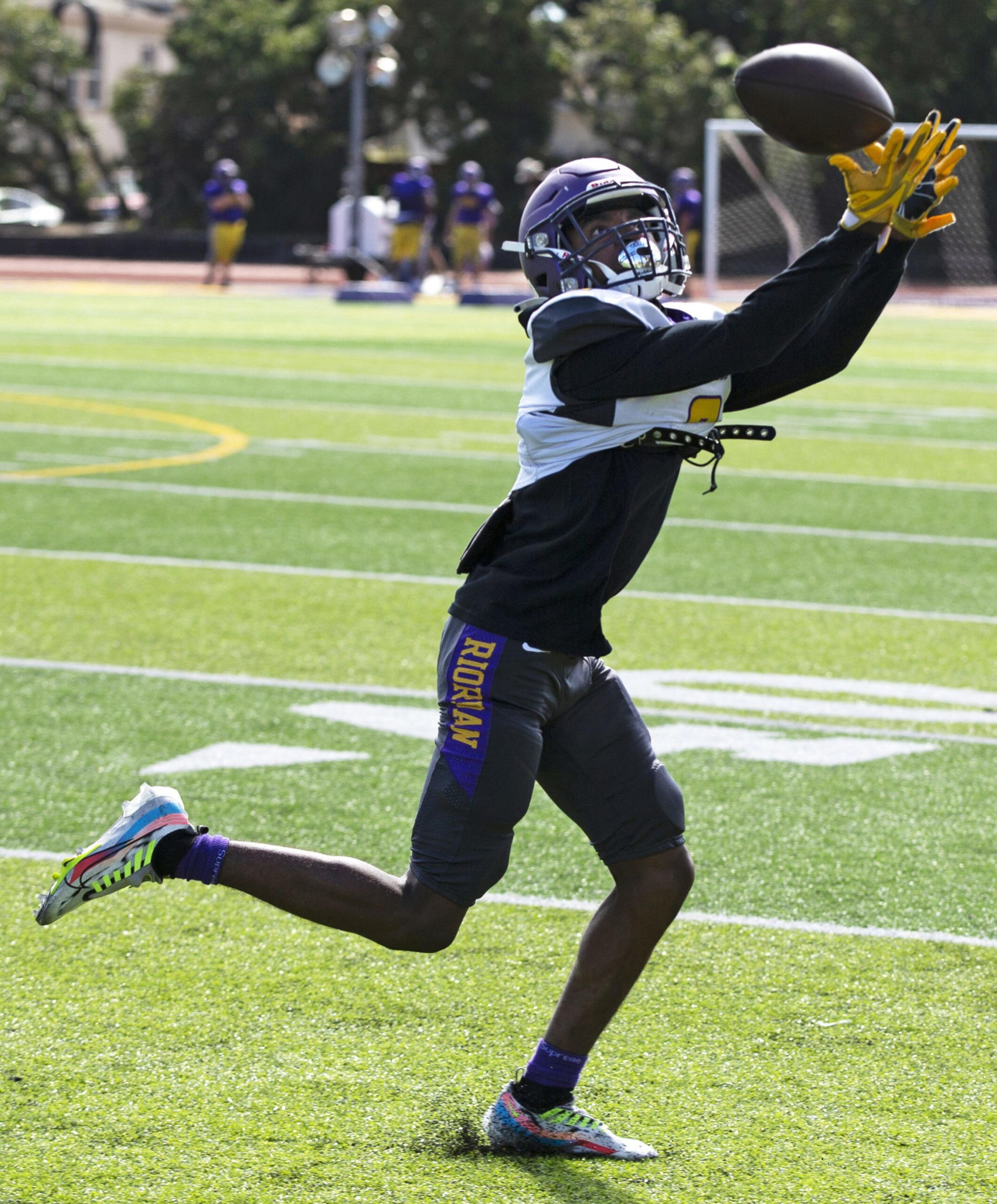 A football player wearing a helmet and yellow gloves leaps forward to catch a football on a sunny green field.