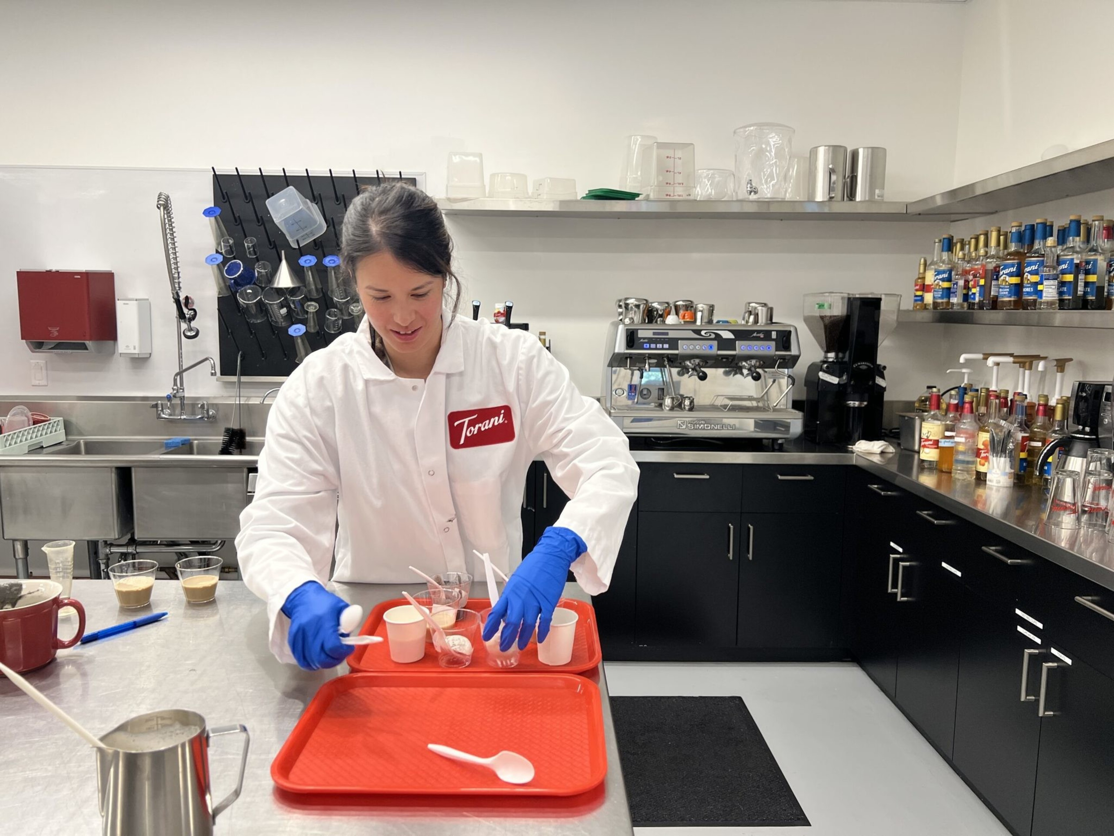 A person in a white coat and blue gloves prepares coffee samples on a red tray in a modern kitchen with coffee machines and syrups.