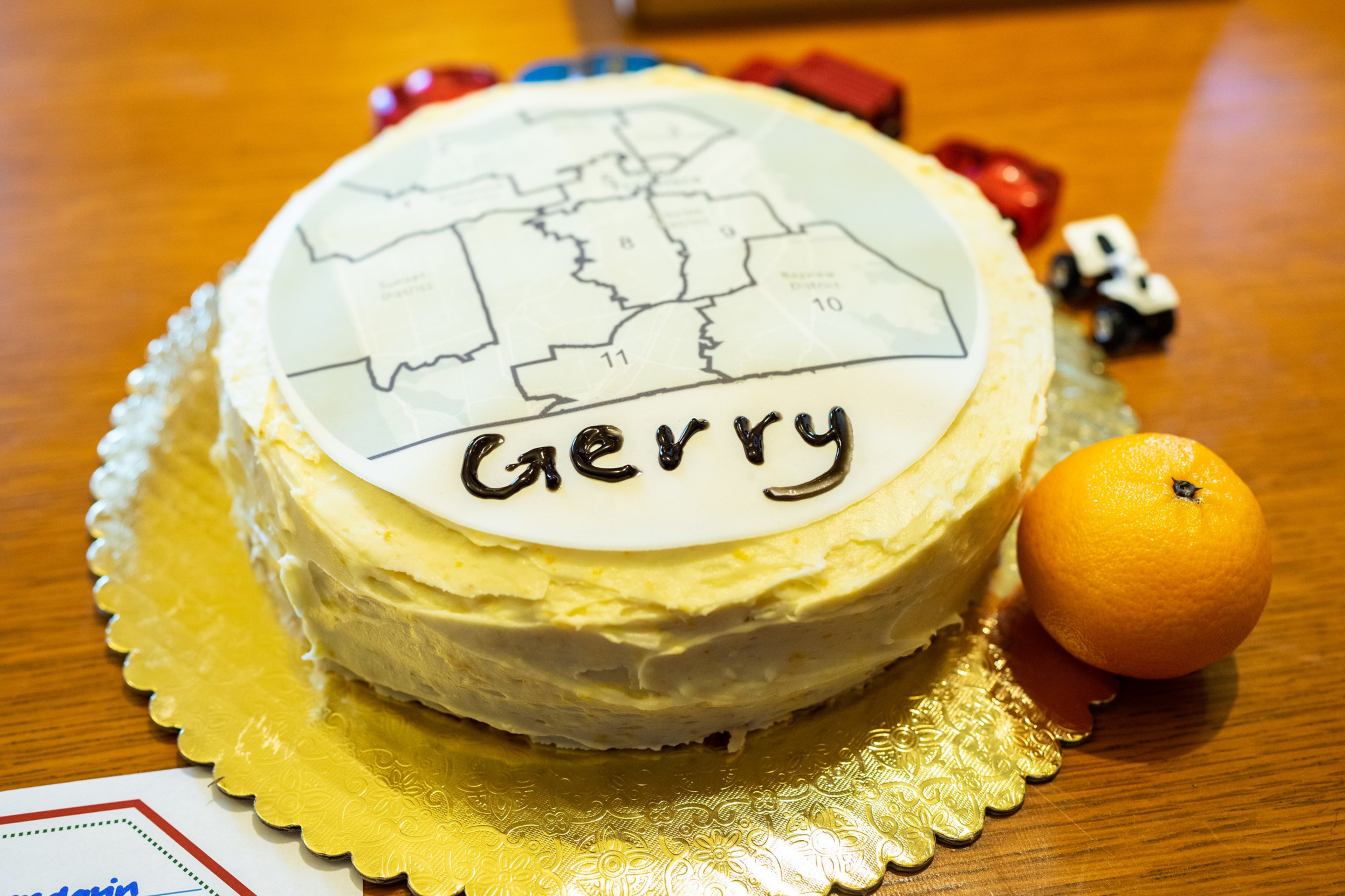 A round cake with white frosting has a map and the word "Gerry" written on top, placed on a golden cake board with a small orange beside it.