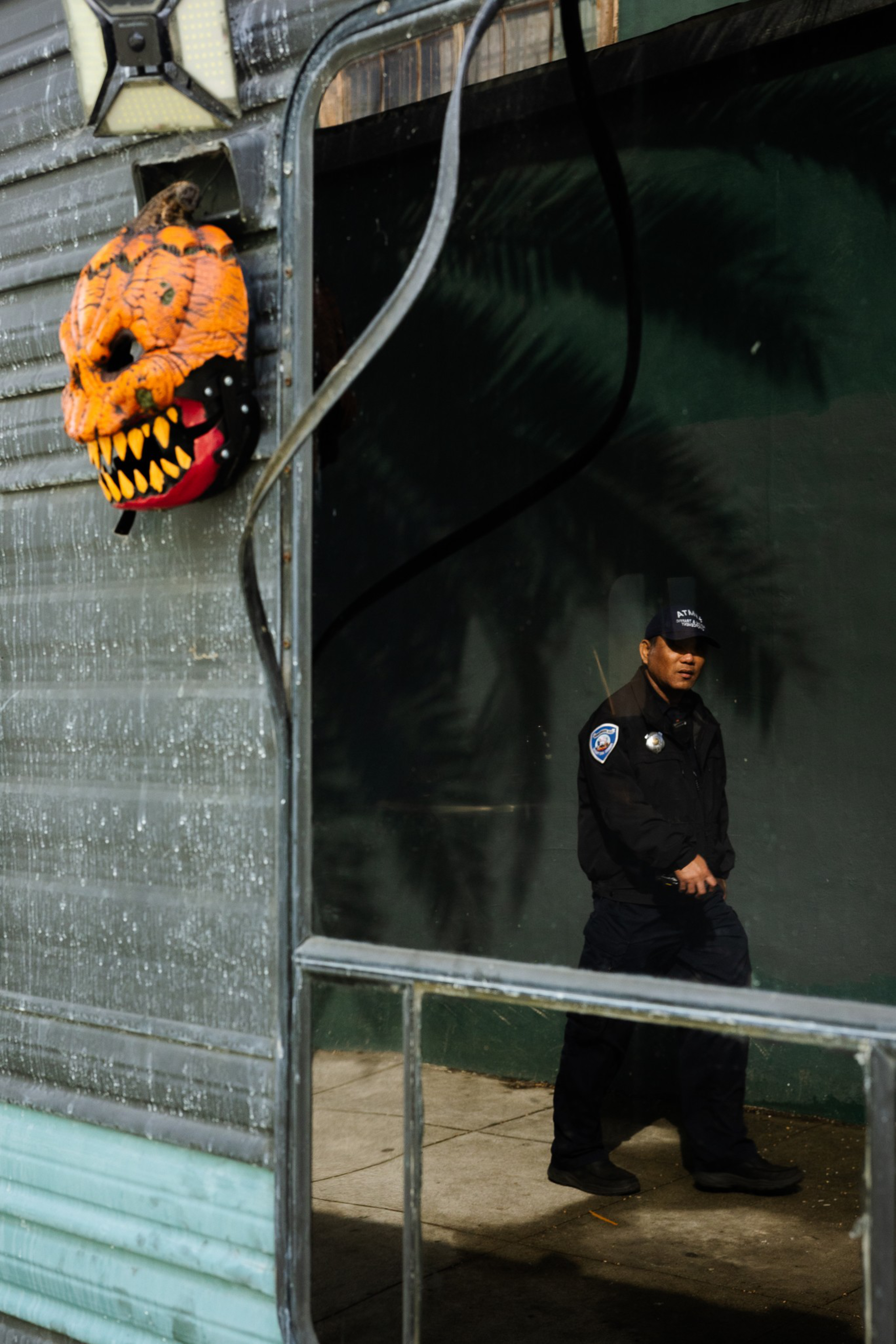 A man in a black uniform and cap walks past a reflective window, beside a wall-mounted orange pumpkin decoration with sharp yellow teeth.