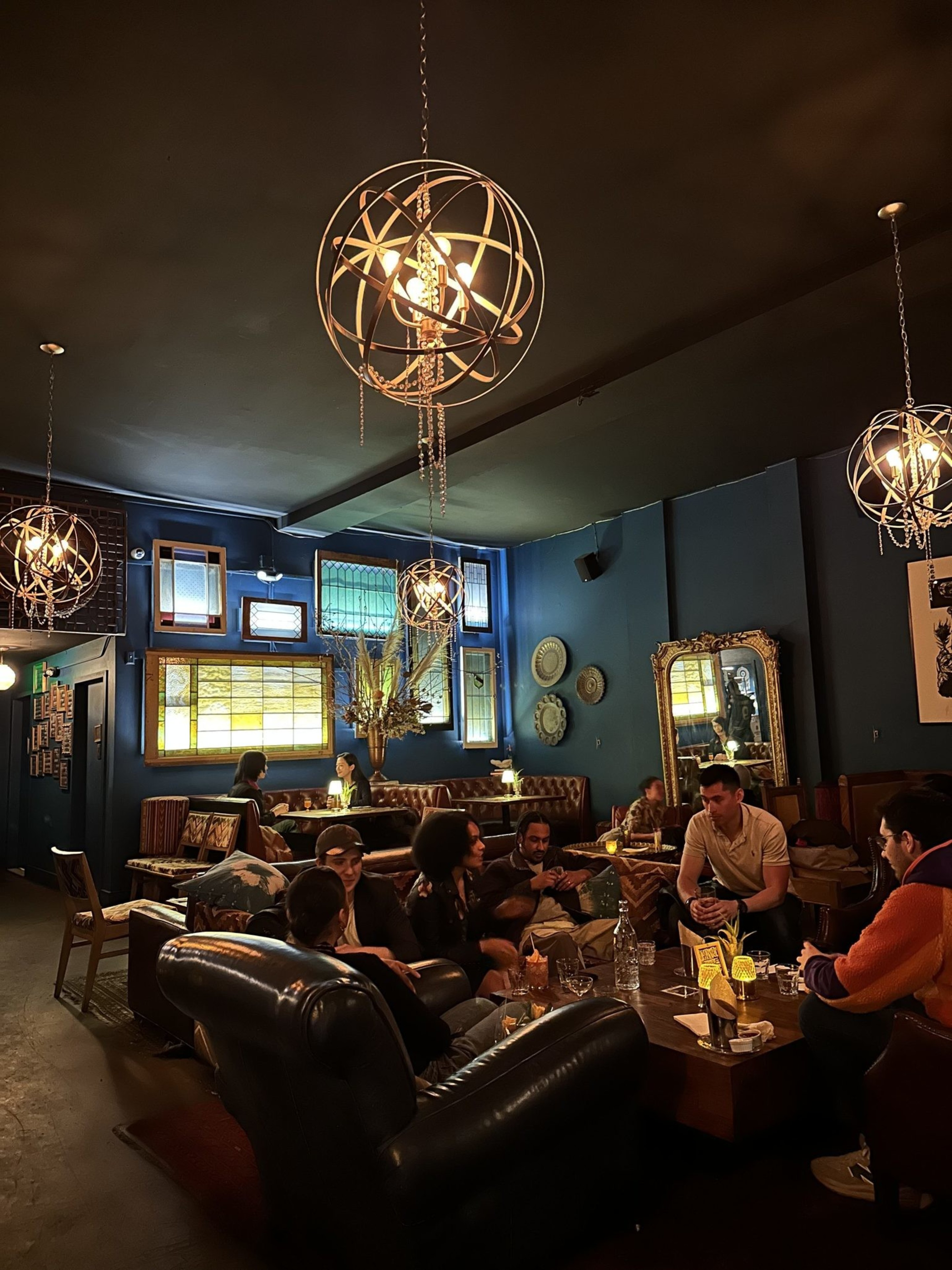 A dimly lit lounge with spherical pendant lights; people sit on leather couches, conversing. The room has blue walls and eclectic decor, creating a cozy atmosphere.