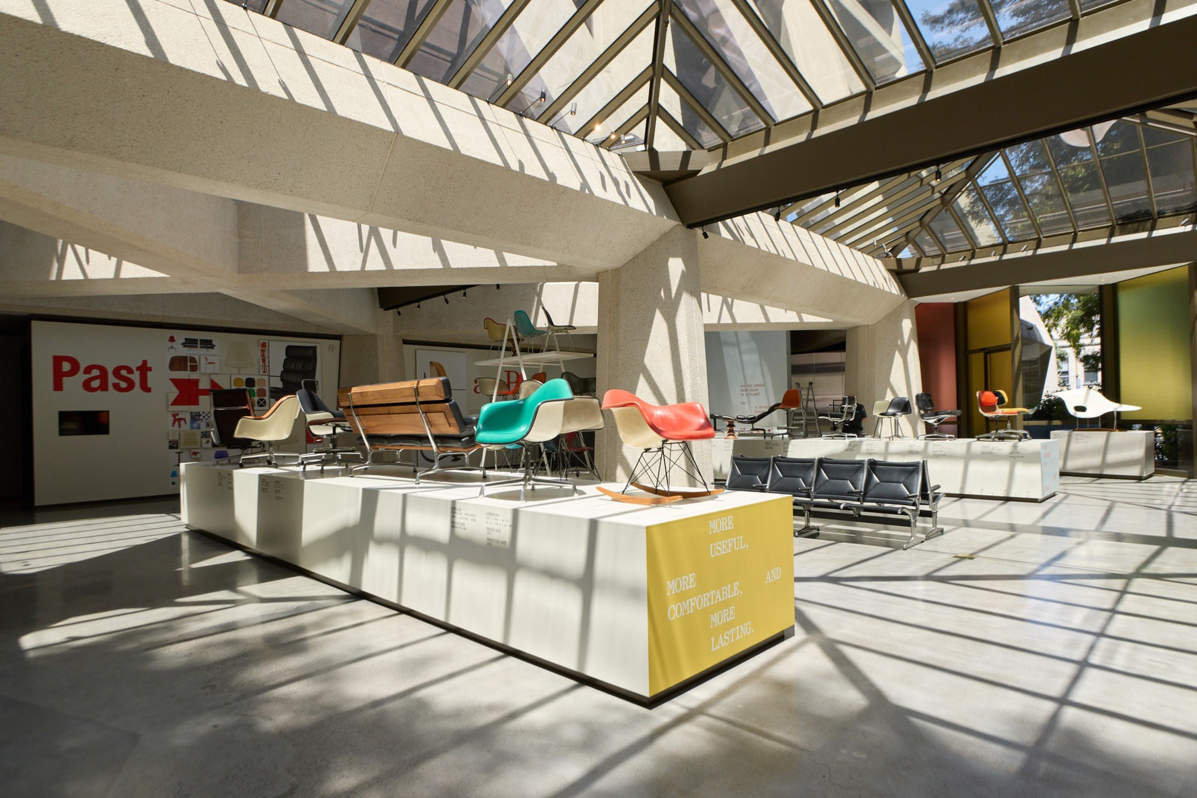 The image shows a sunlit exhibition space with modern chairs displayed on platforms. There's a large &quot;Past&quot; sign and colorful walls, creating a bright, airy atmosphere.