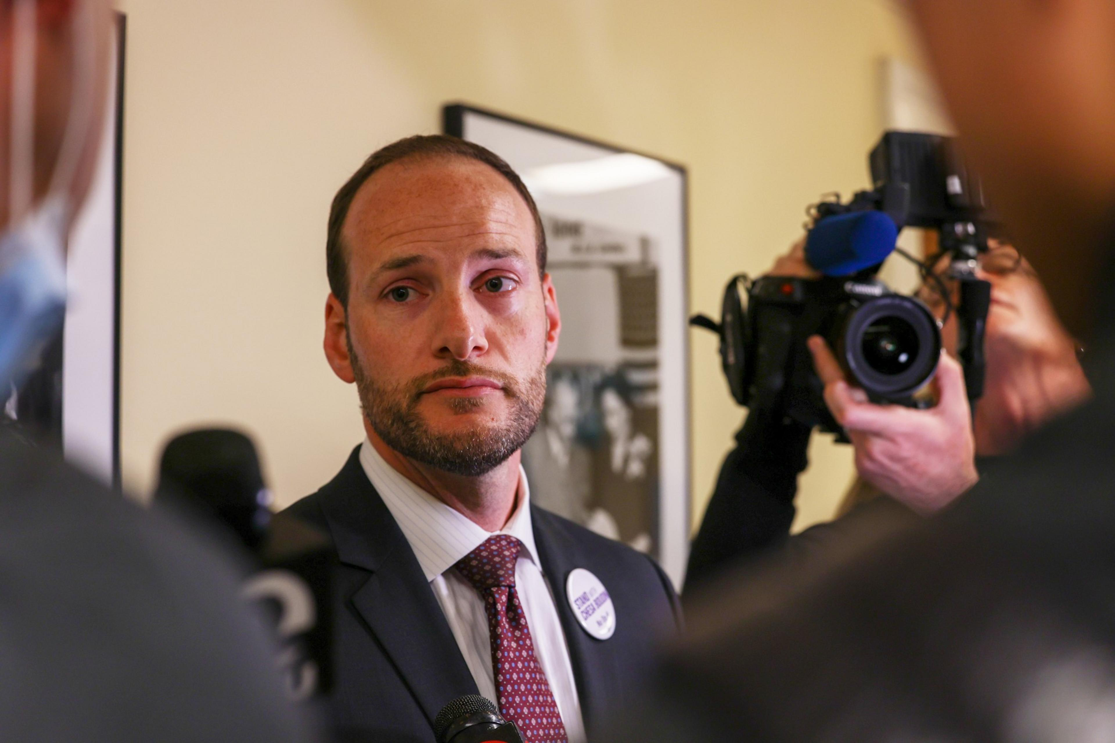 A man in a suit and tie with a campaign button looks solemn while being interviewed, with a camera and microphone pointed at him.