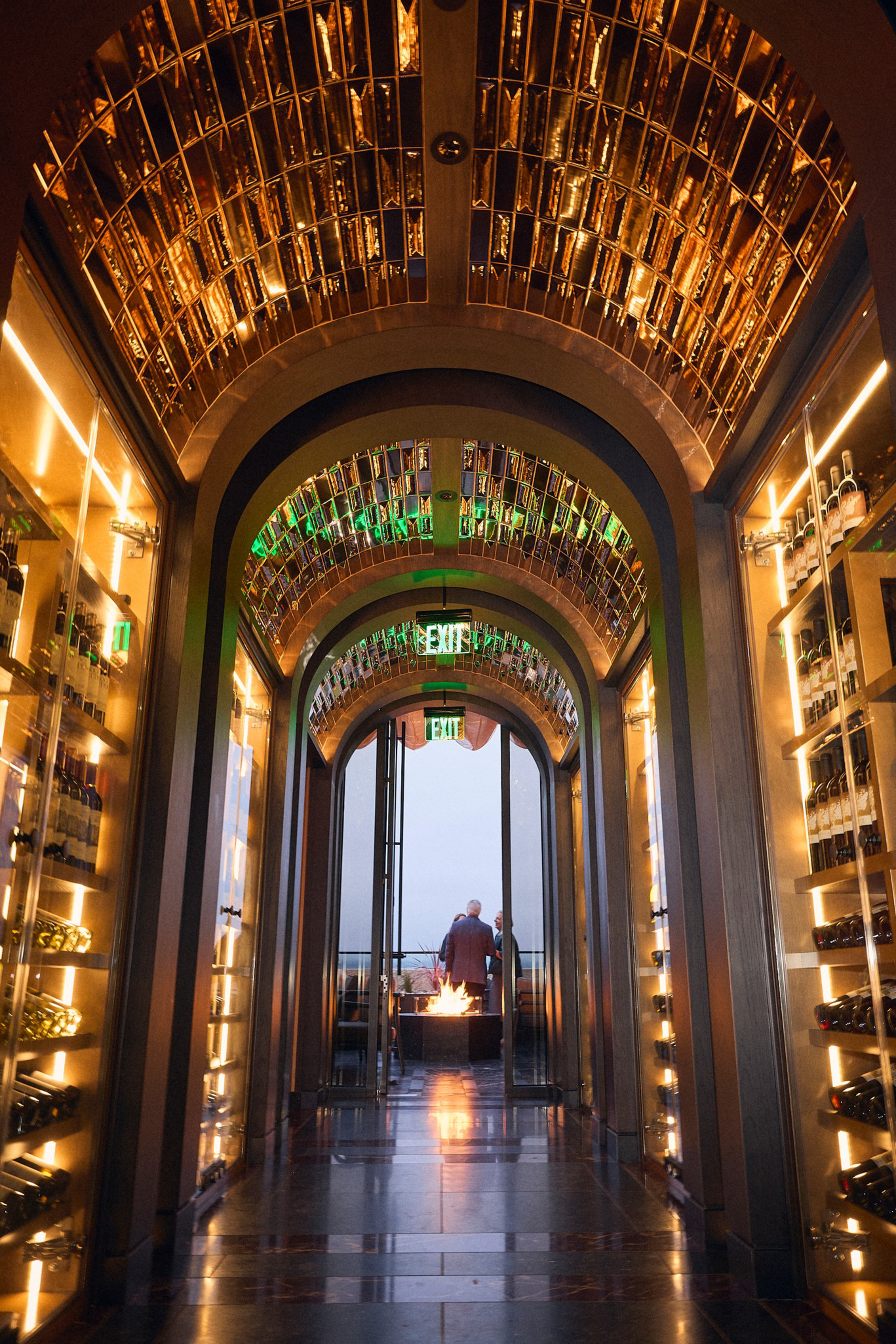A lit arched hallway lined with illuminated wine racks leads to an open door, where two people stand by a fire pit outside.