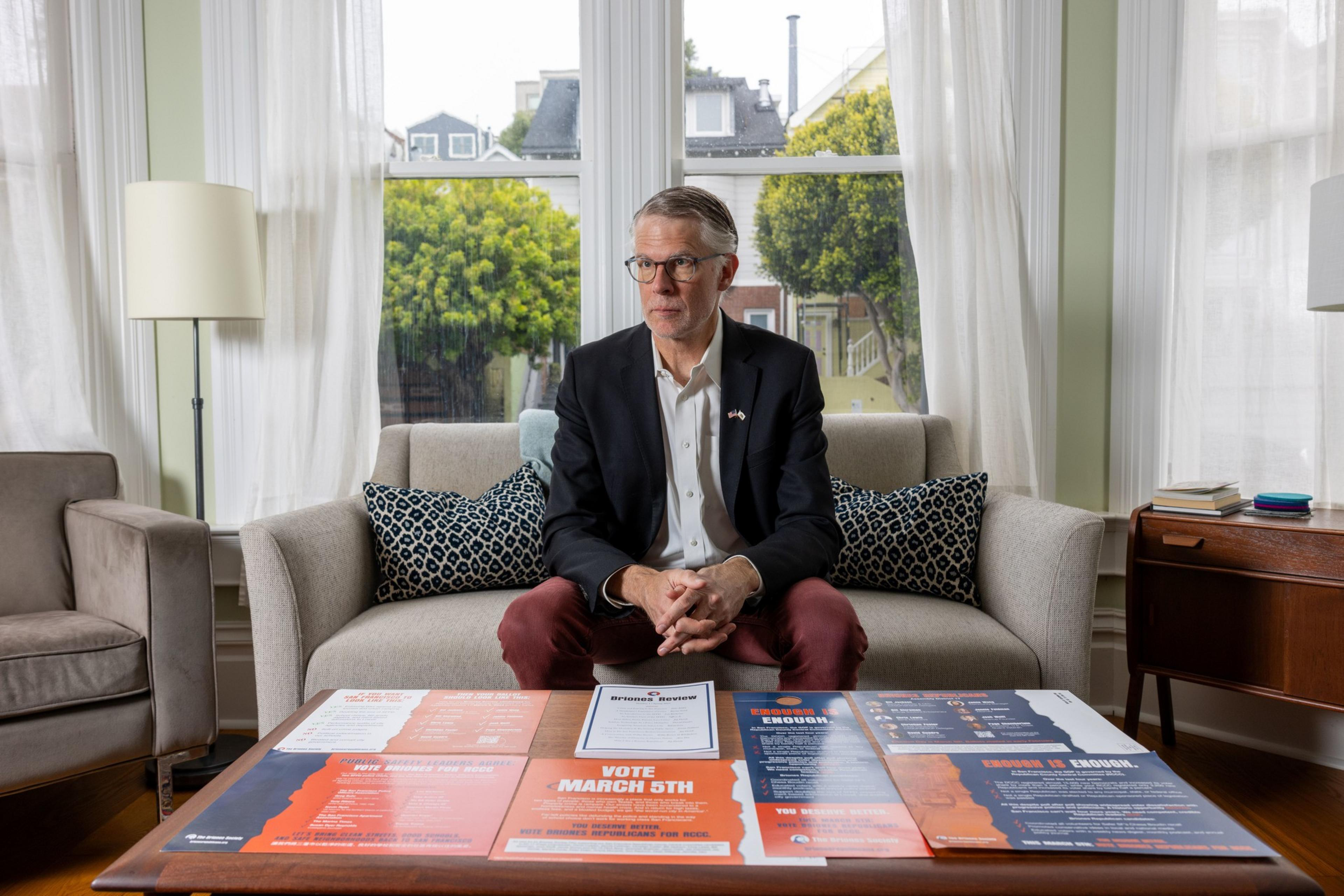 A man in a dark suit sits on a beige sofa in a well-lit living room, with campaign posters spread out on the coffee table in front of him.