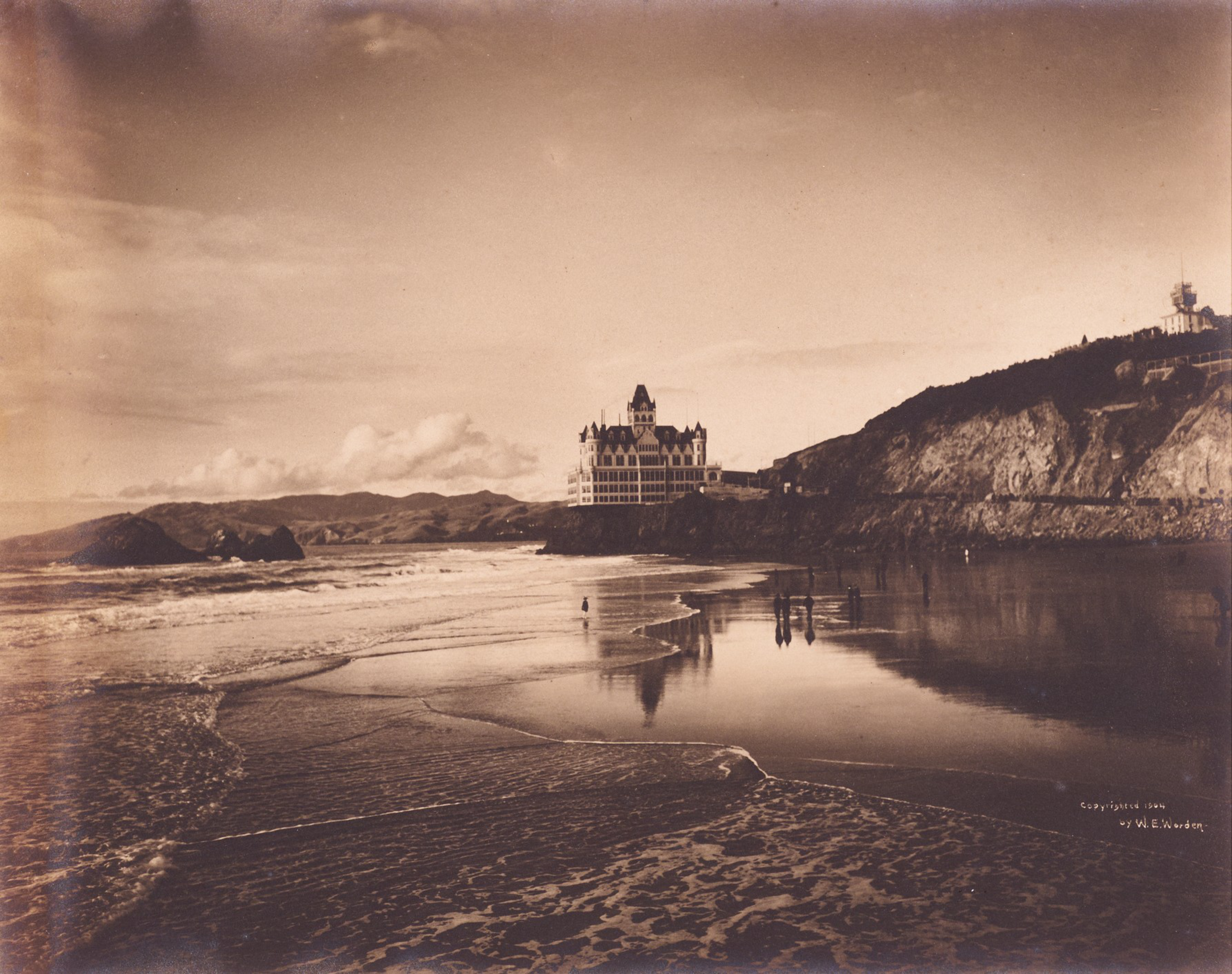 A castle-like building stands on a cliff overlooking a beach with gentle waves and a few people walking along the wet sand, reflecting the sky.