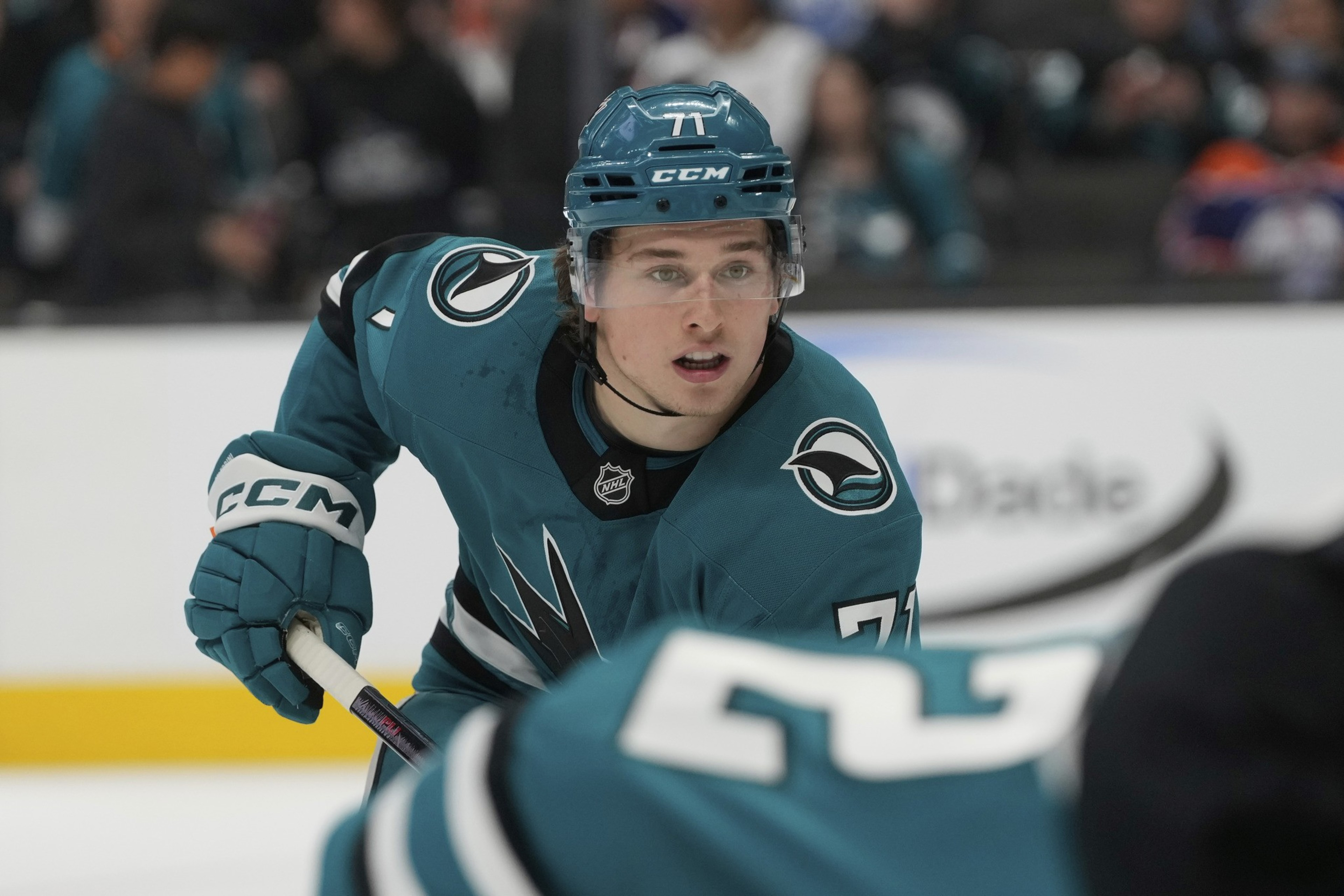A hockey player wearing a teal jersey and helmet skates on the ice, gripping a stick. Spectators blur in the background.