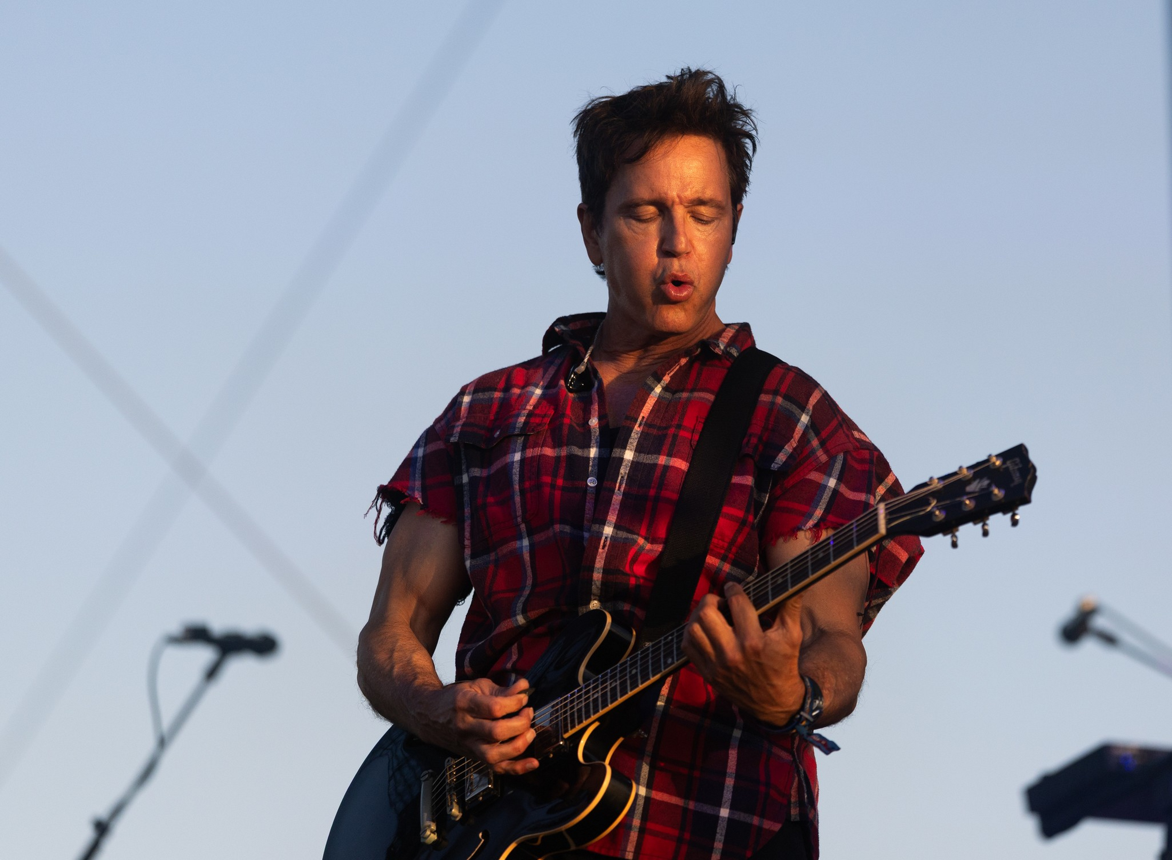 A man wearing a red plaid sleeveless shirt plays an electric guitar with eyes closed and lips pursed, performing on an outdoor stage.