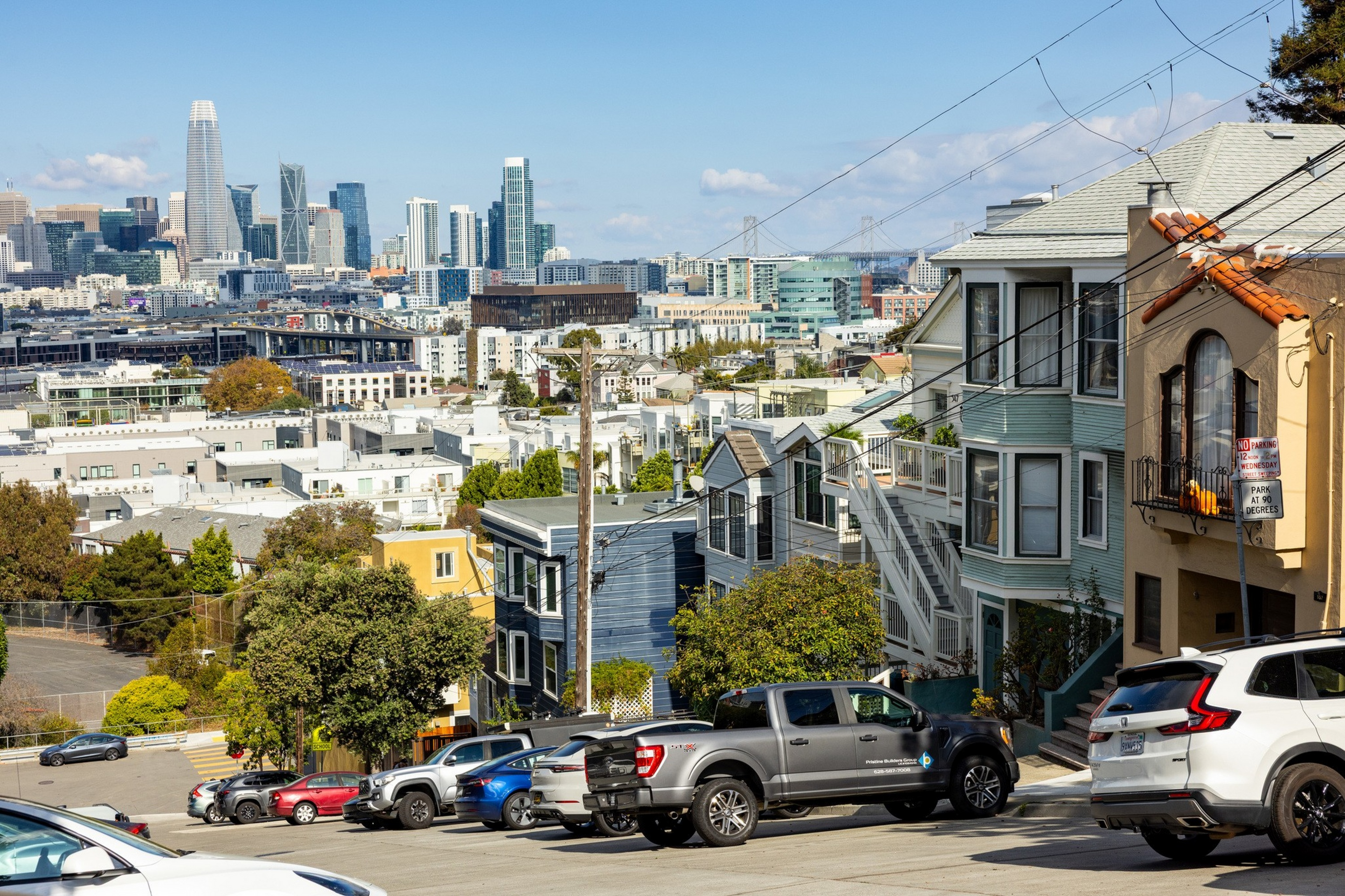 A steep city street lined with parked cars and colorful houses slopes downward toward a downtown skyline with tall modern buildings under a blue sky.