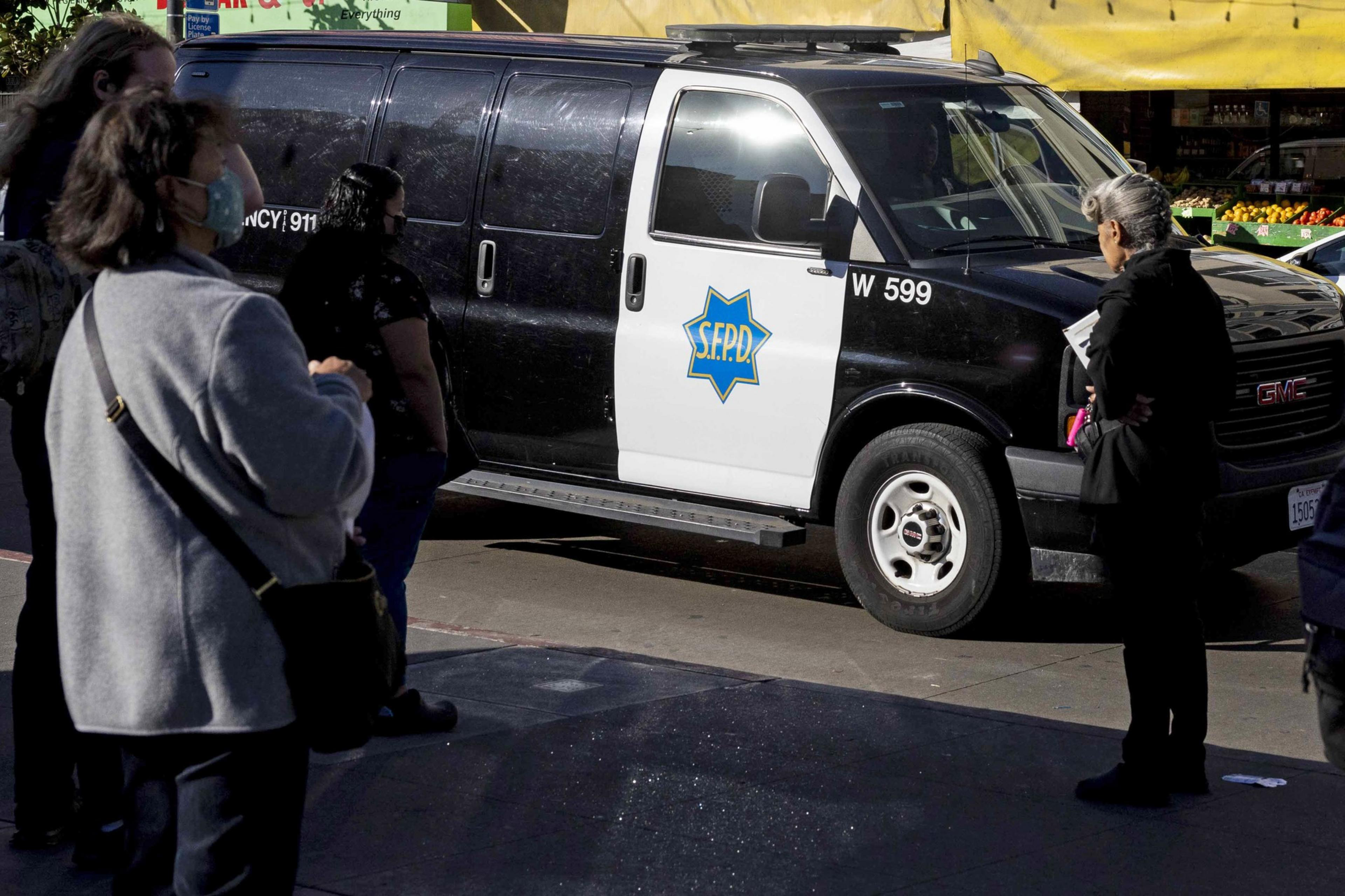 A black and white SFPD van is parked on the street. People are standing nearby, some wearing masks. Thereâ€™s a market stall in the background.