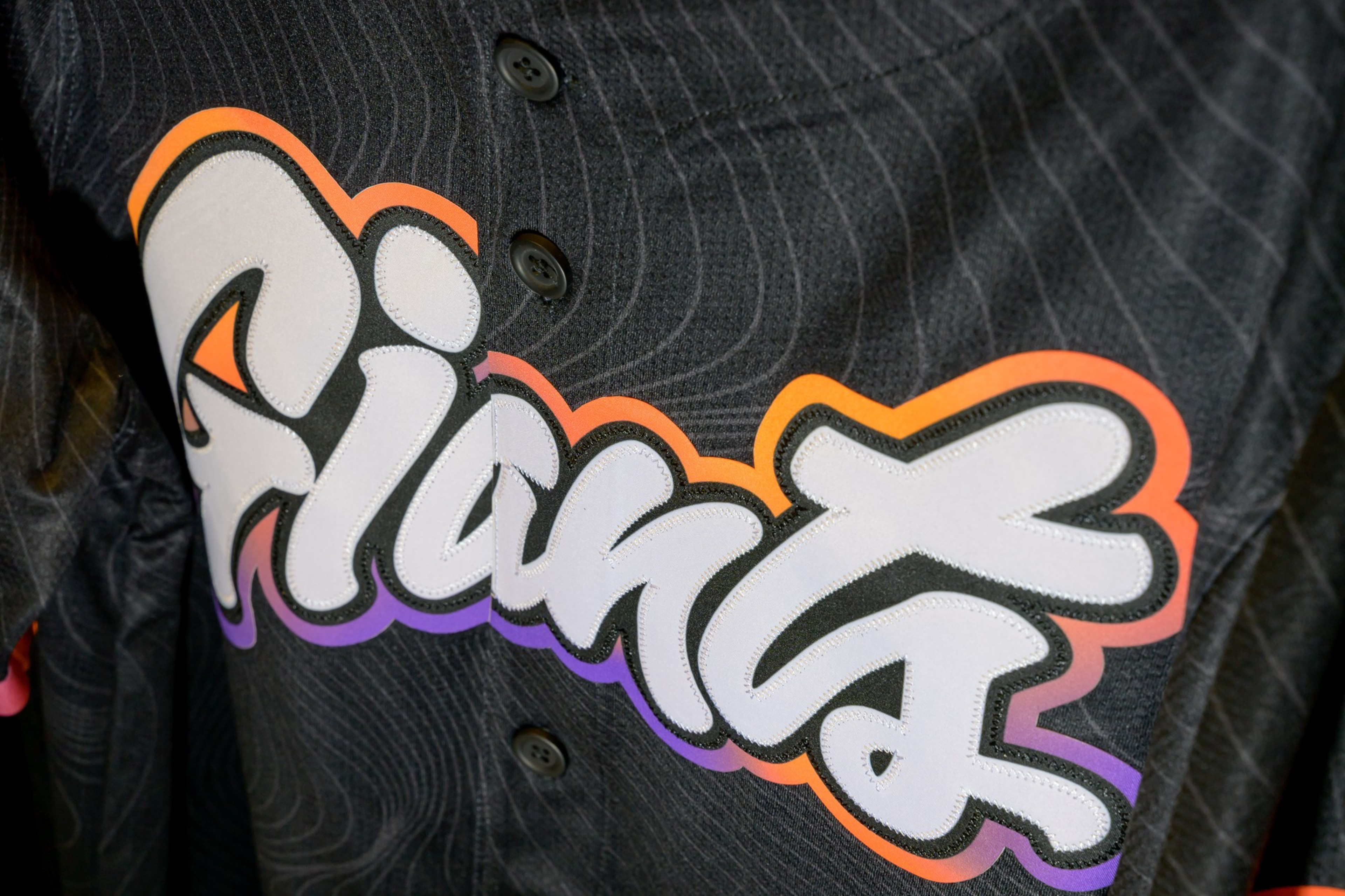 The image shows a close-up of a black jersey with &quot;Giants&quot; written in bold, playful letters, outlined in orange and purple. The jersey has black buttons.
