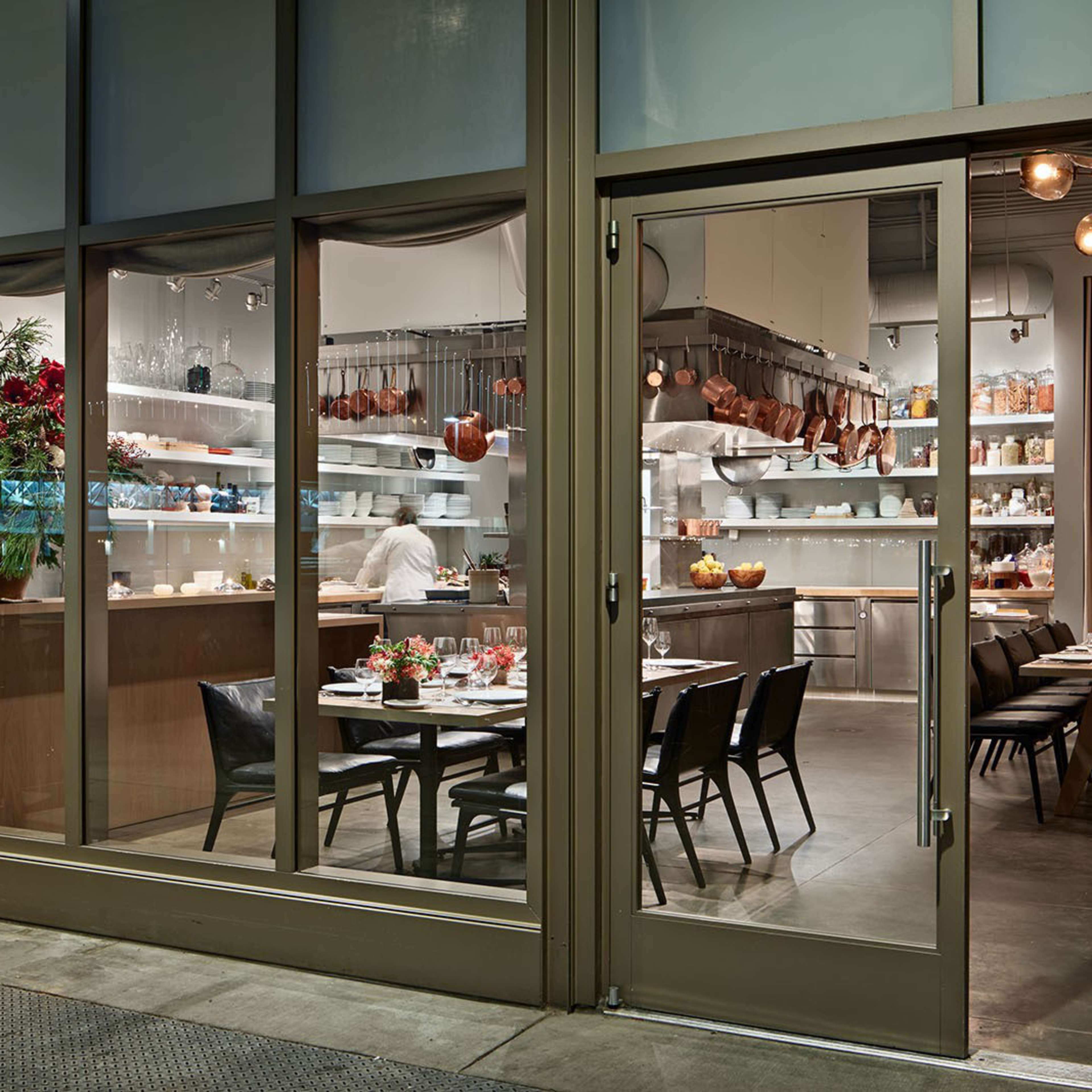 A modern dining area with two set tables, black chairs, a well-organized kitchen with copper pots, open shelves, and a glass entrance door.