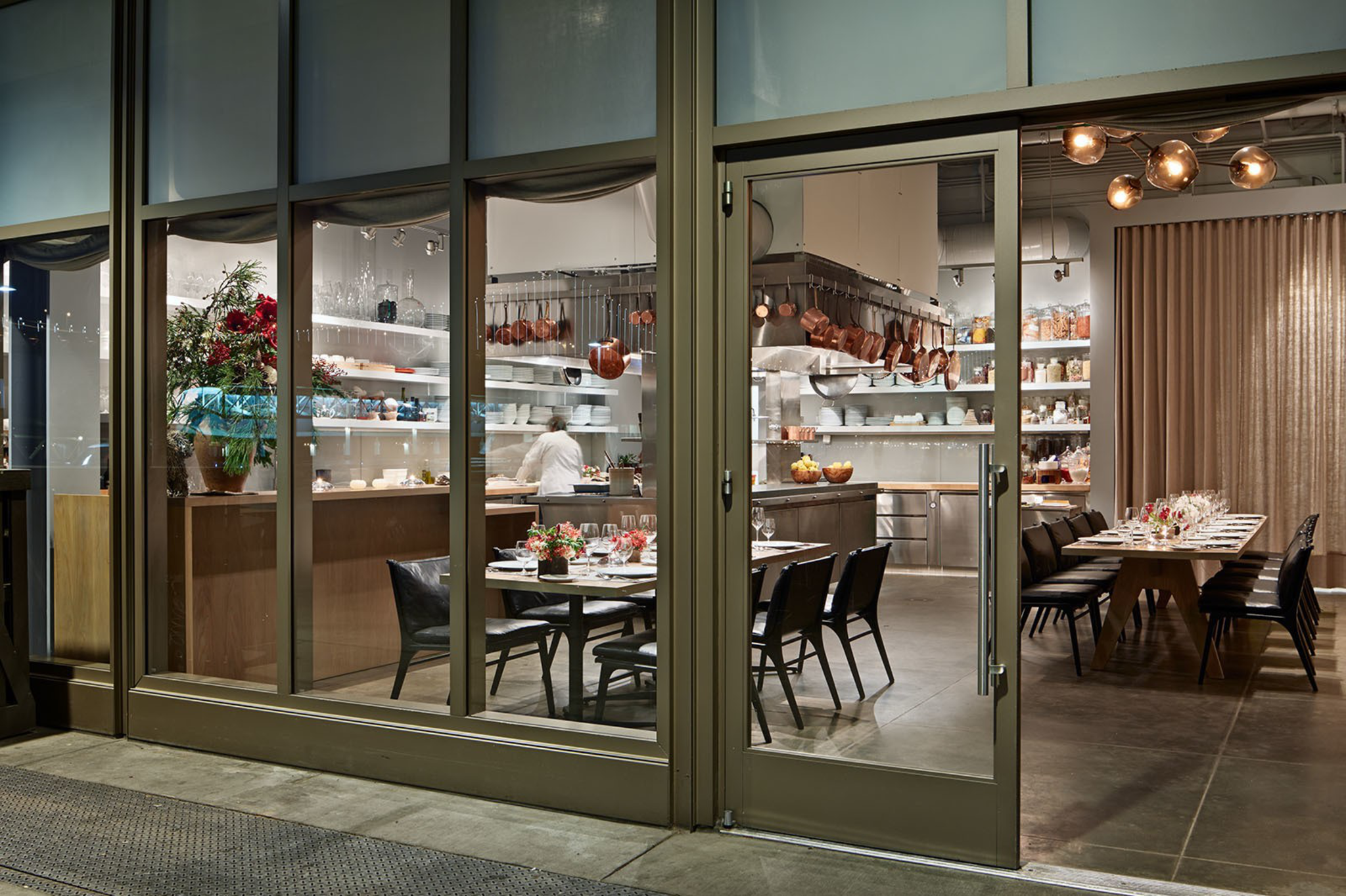 A modern dining area with two set tables, black chairs, a well-organized kitchen with copper pots, open shelves, and a glass entrance door.