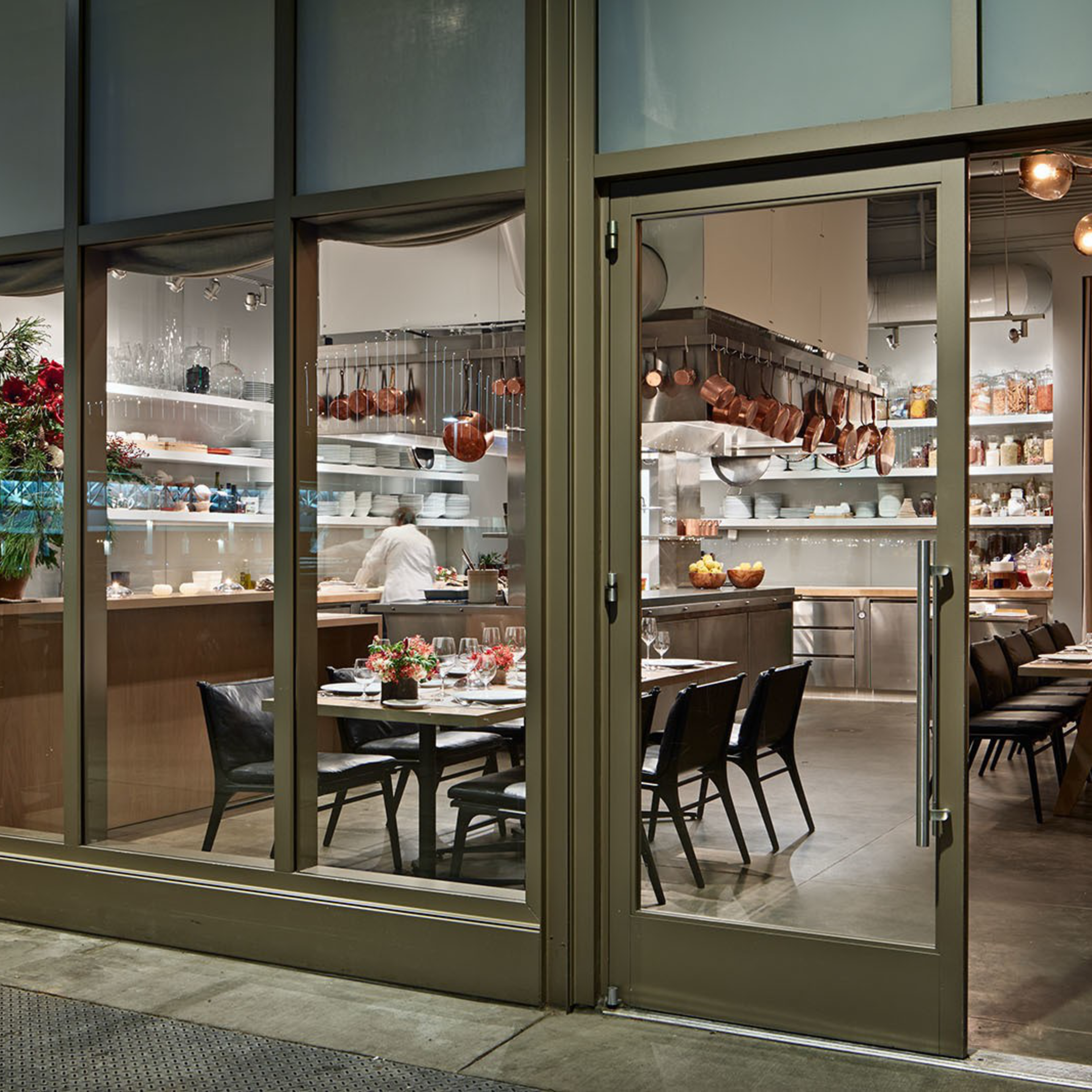 A modern dining area with two set tables, black chairs, a well-organized kitchen with copper pots, open shelves, and a glass entrance door.