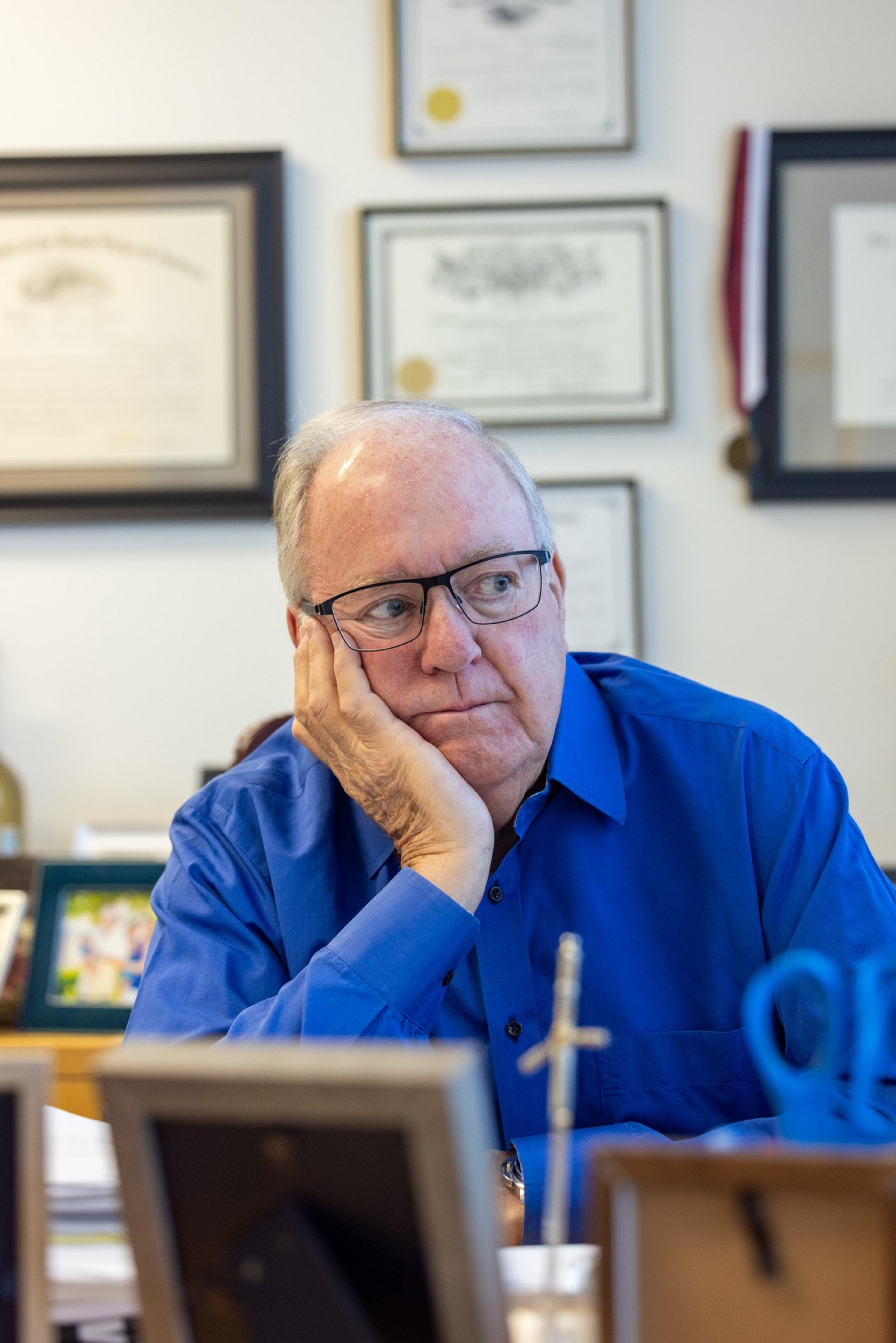 A man in glasses and a blue shirt rests his head on his hand, looking contemplatively. Several framed certificates and photos are visible on the wall behind him.