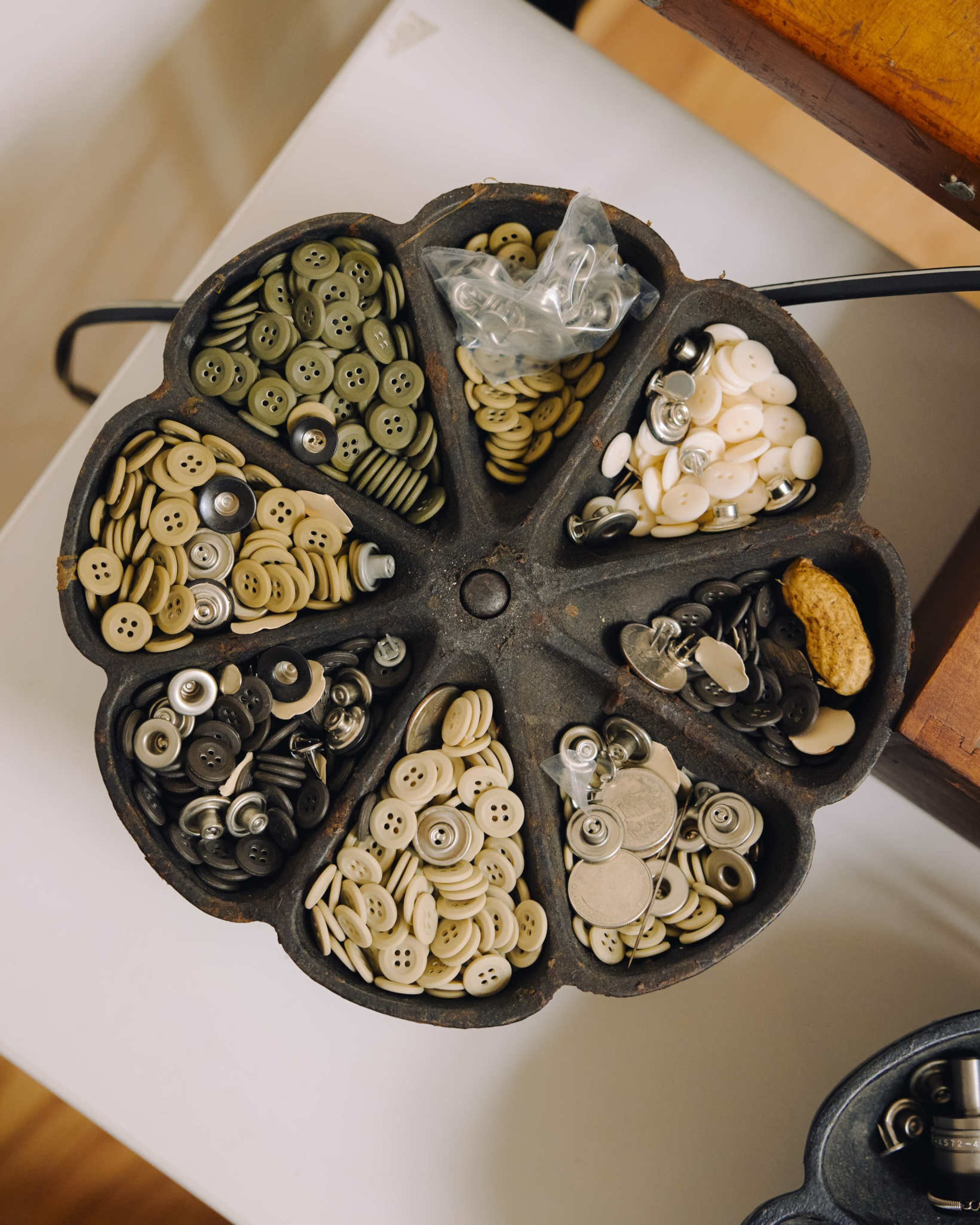 A round divided metal tray holds various buttons in different colors and sizes, with a peanut resting in one section.