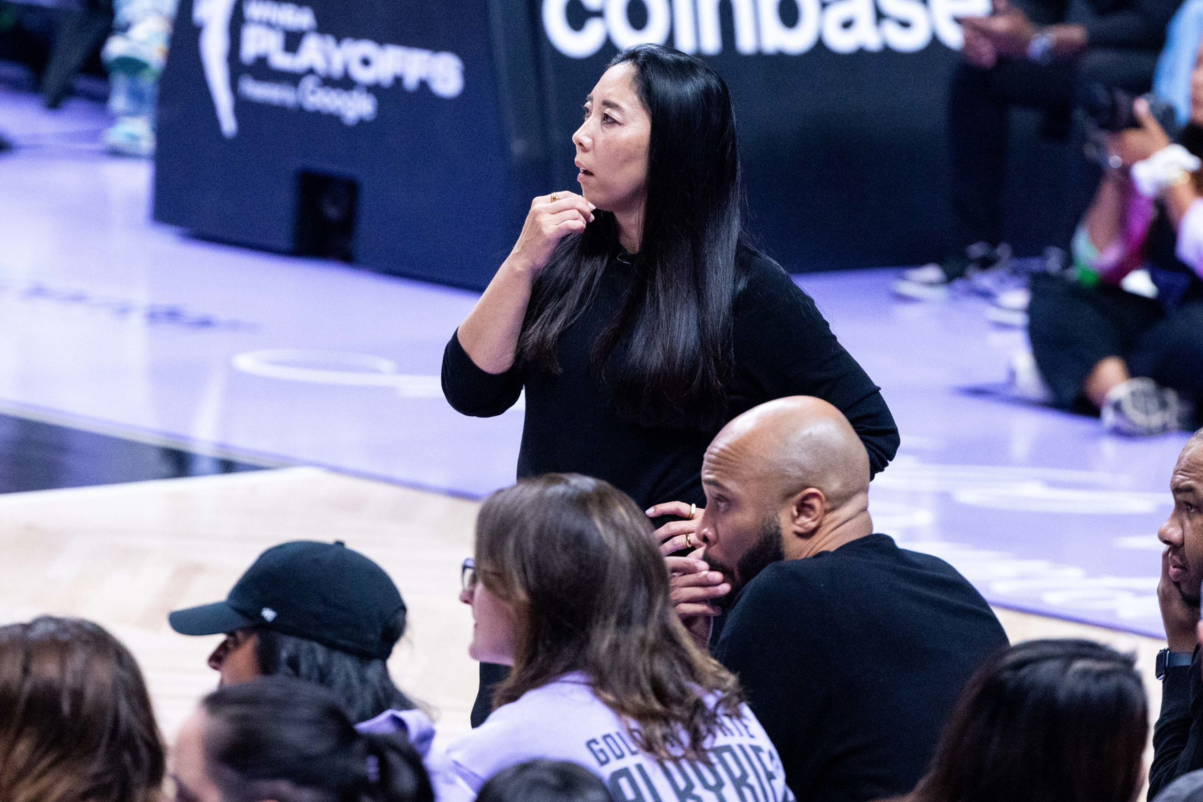 A woman with long black hair stands near a basketball court, looking focused, while people in front of her sit and watch the game.