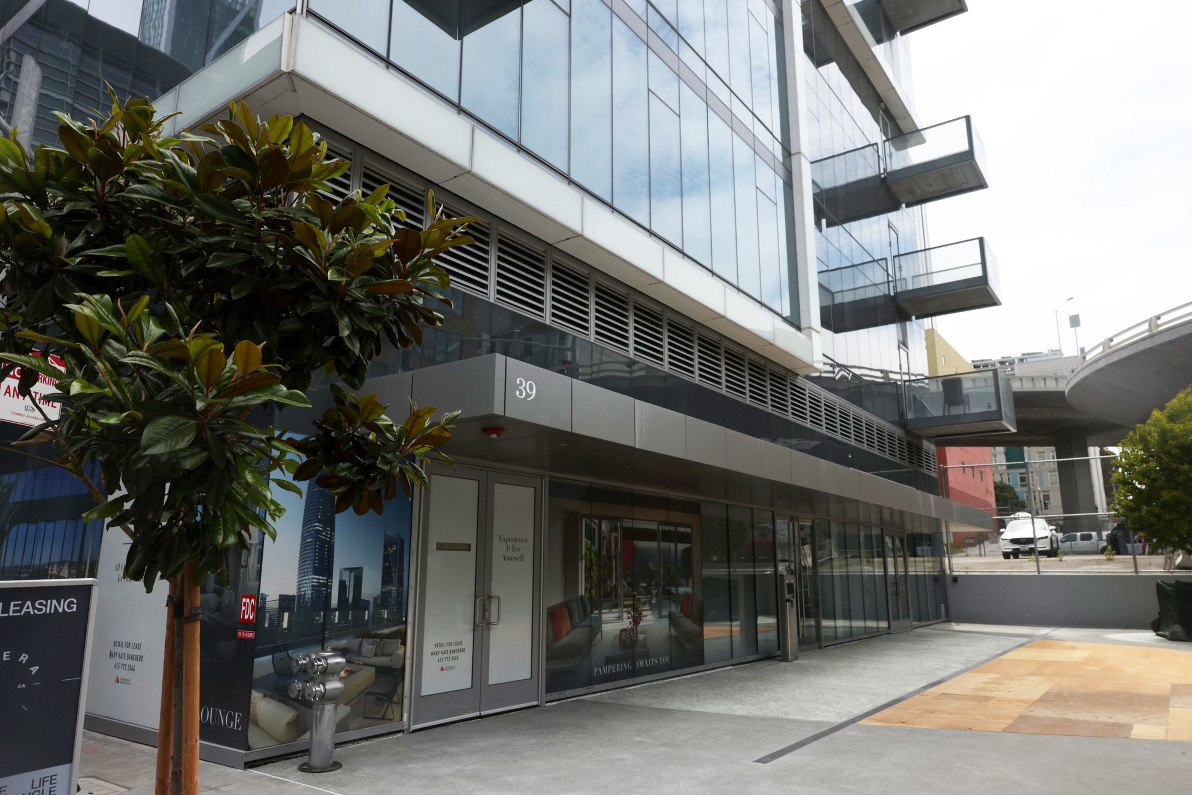 The image features a modern glass building exterior with balconies, a "39" sign, and a leasing advertisement. A young tree is in the foreground near a clean, paved walkway.