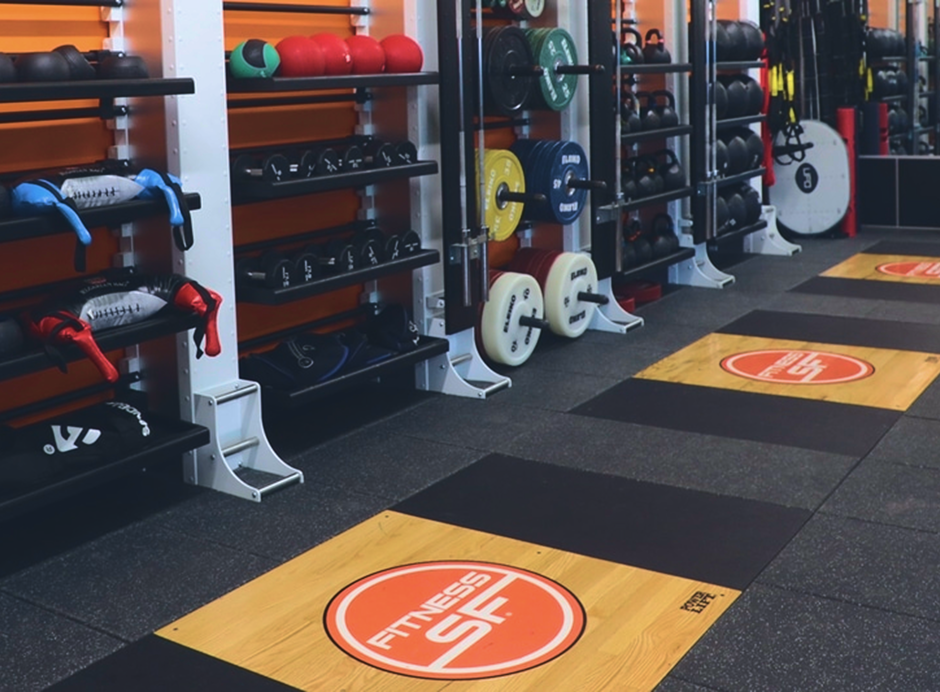 Weightlifting platforms with "Fitness SF" logos are lined along a gym wall filled with dumbbells, medicine balls, and weight plates.